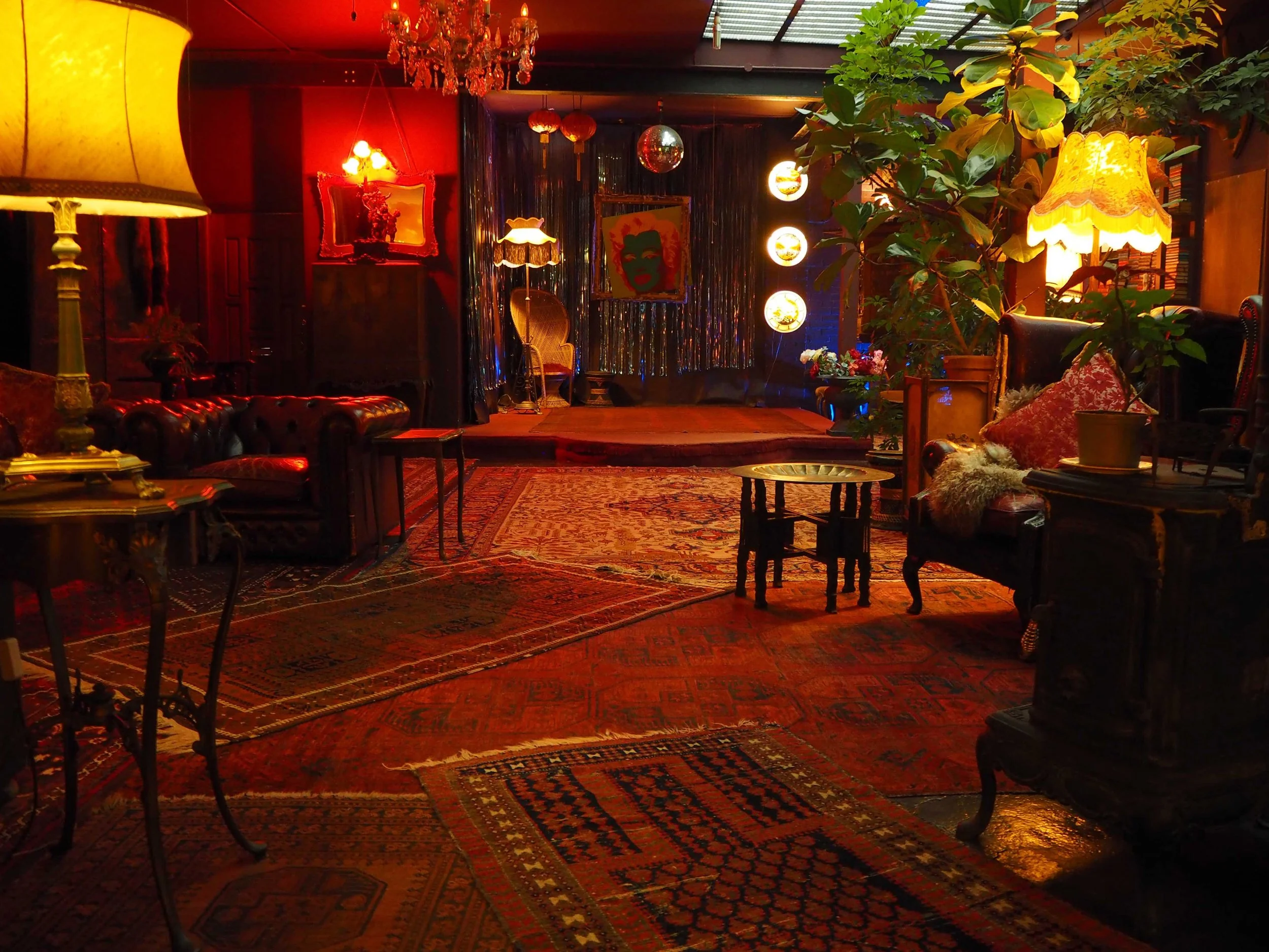 A cozy lounge with vintage furniture, warm lighting, rugs, and a small stage decorated with curtains, artwork, and ornamental lighting.