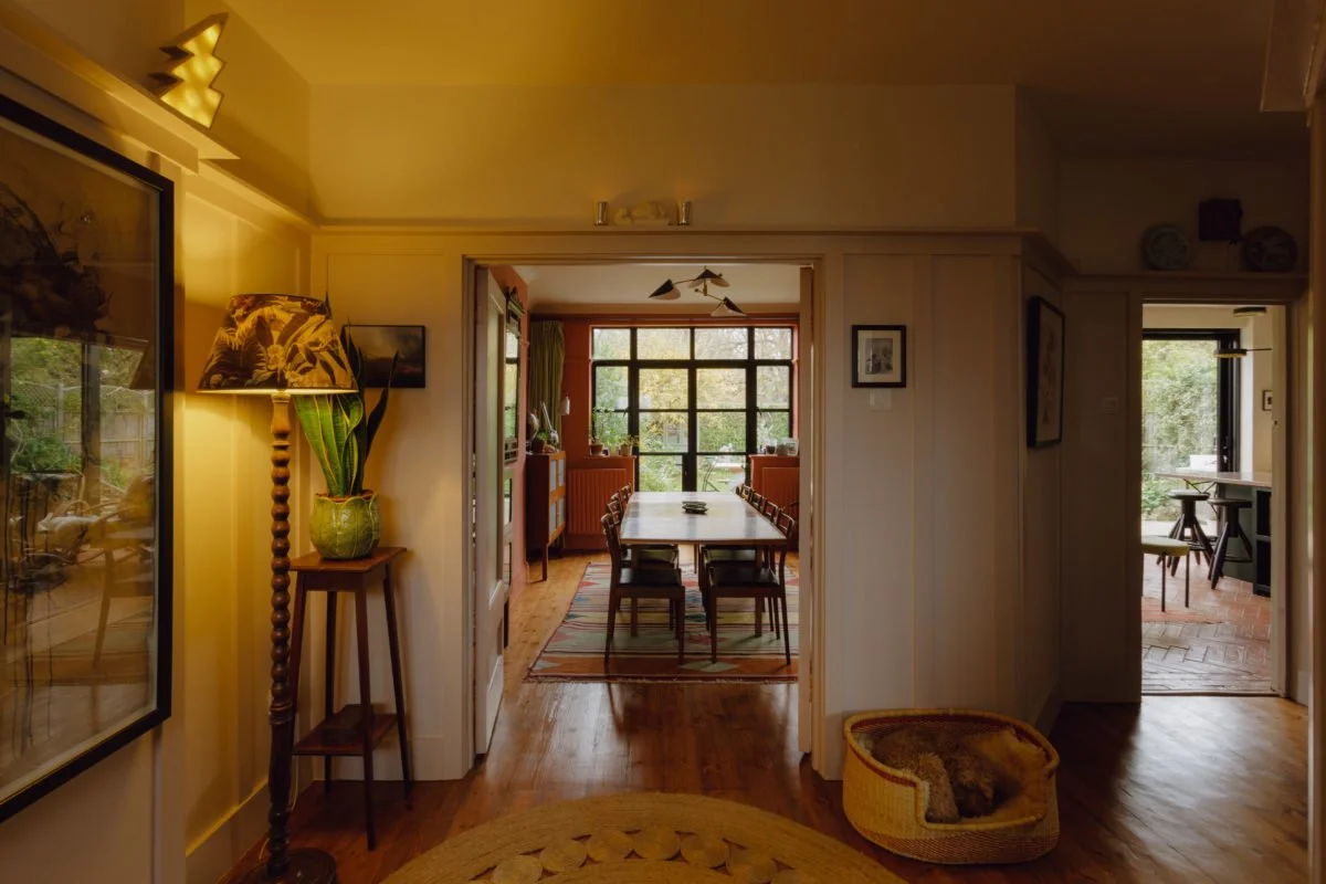 Interior view of a dining area with a long wooden table and chairs, leading to a sunroom with large windows, and a side room with a small table and chairs, decorated with plants and artwork.