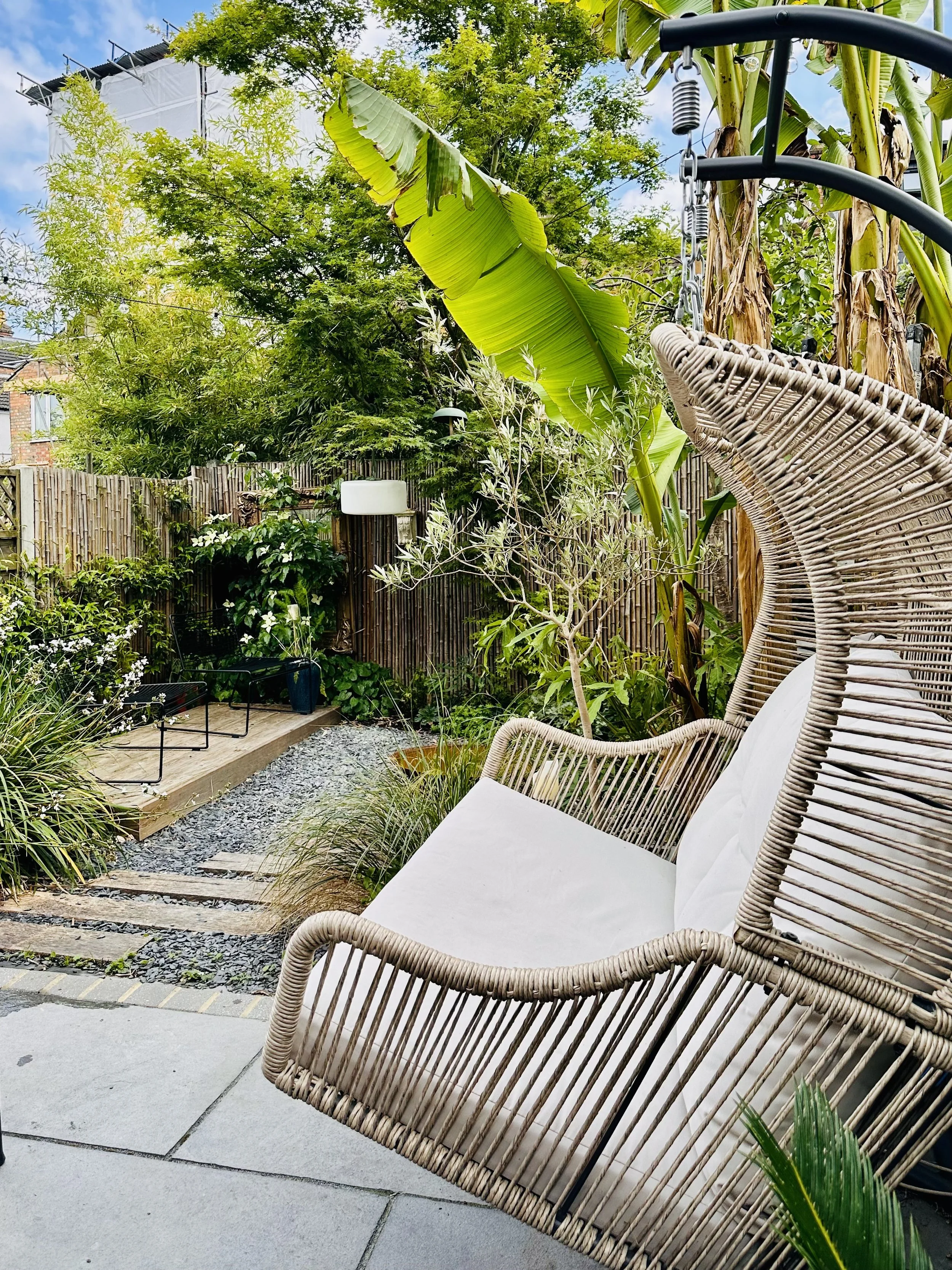 Cozy backyard garden with rattan hanging chair and lush green plants, including banana trees, set against a wooden fence and a partly cloudy sky.