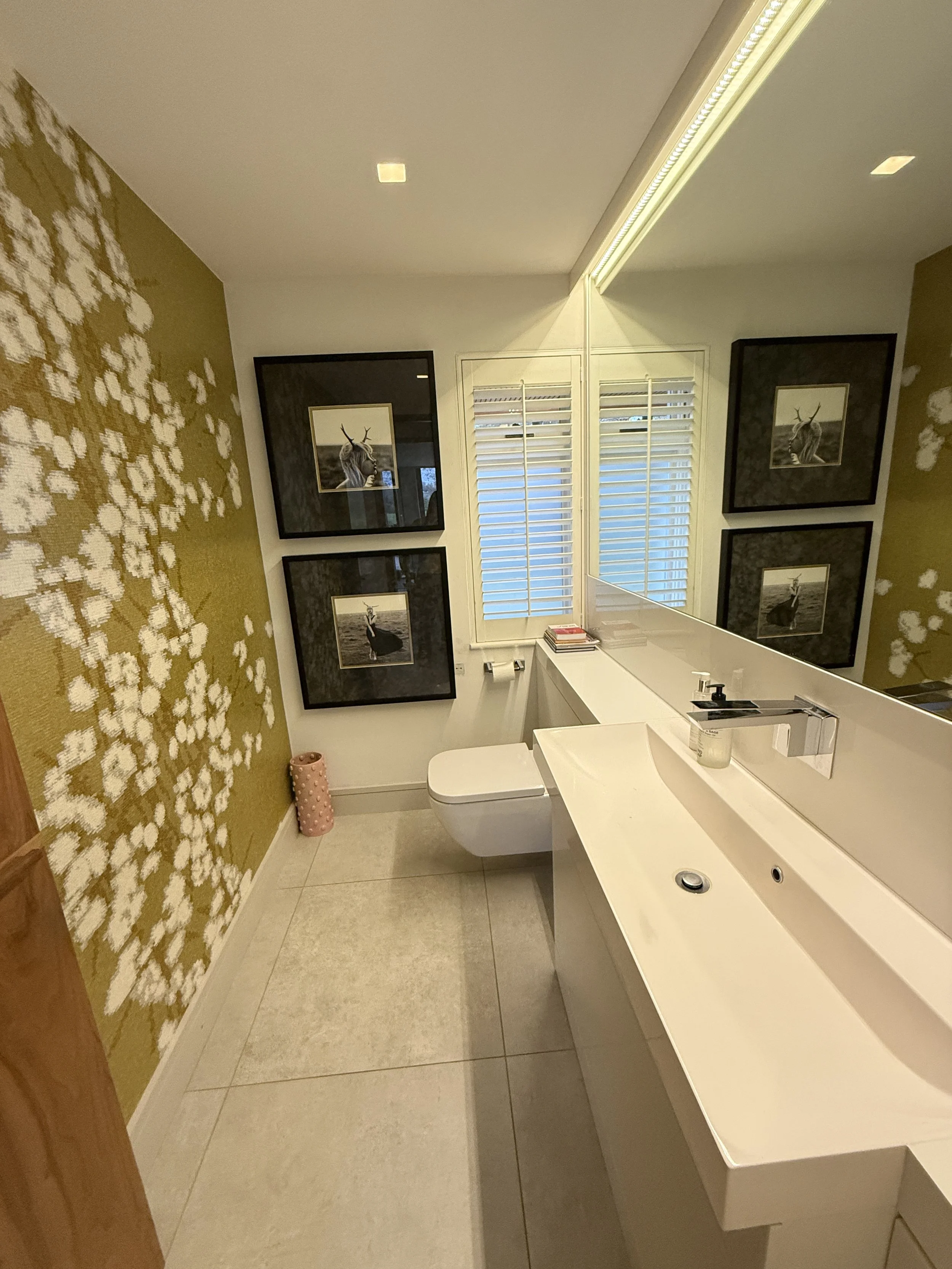 Modern bathroom with a white sink, a toilet, framed black-and-white animal photographs, a mirror, window blinds, a gold-and-white decorative wall, and a pink cylindrical object.