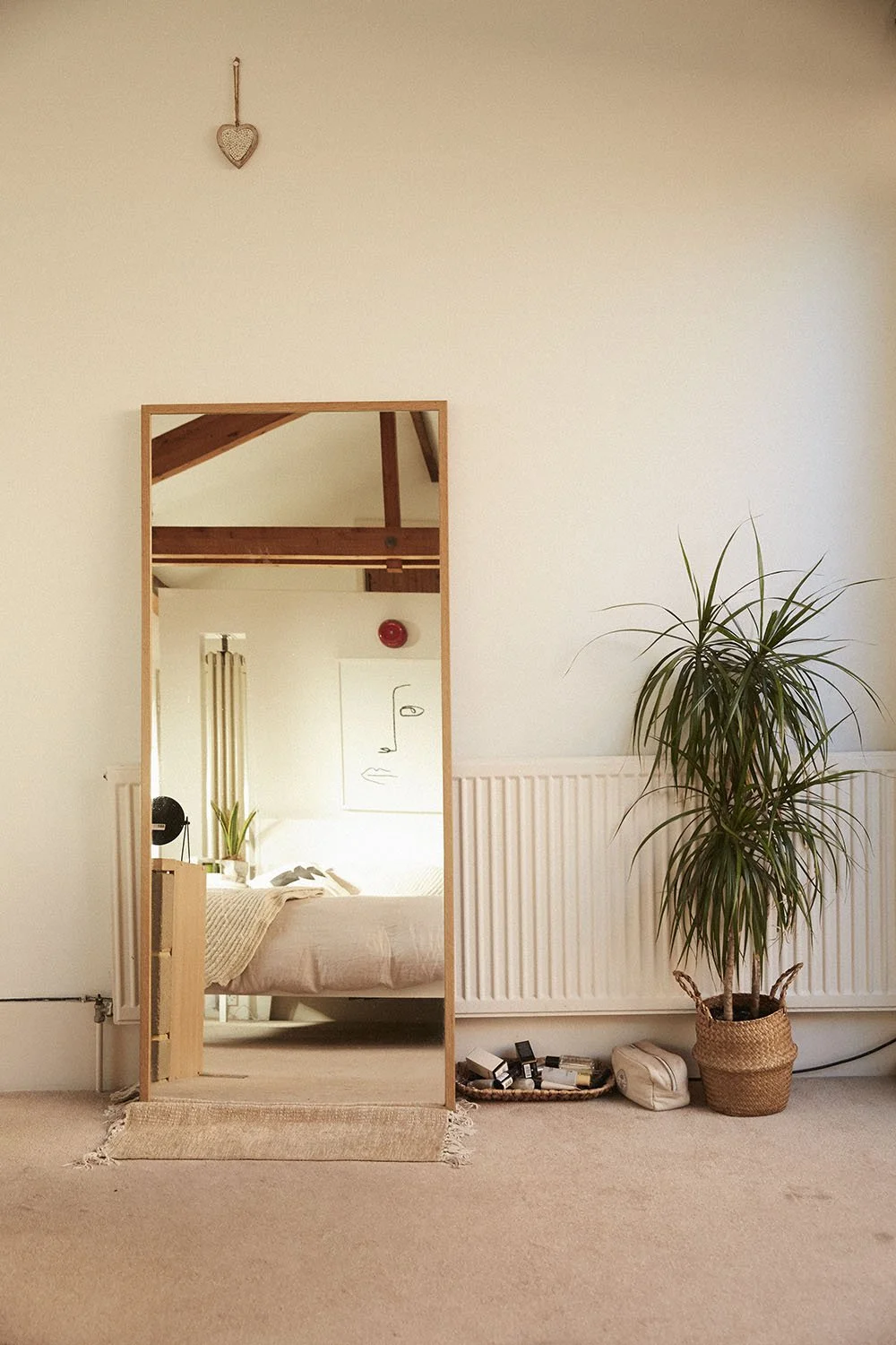A large mirror reflects a bedroom with a bed, a small plant, a lamp, and wall art. Next to the mirror on the right is a tall potted plant, and below it, a small collection of bottles, a bag, and a tray are on the floor. The wall is white with a radia