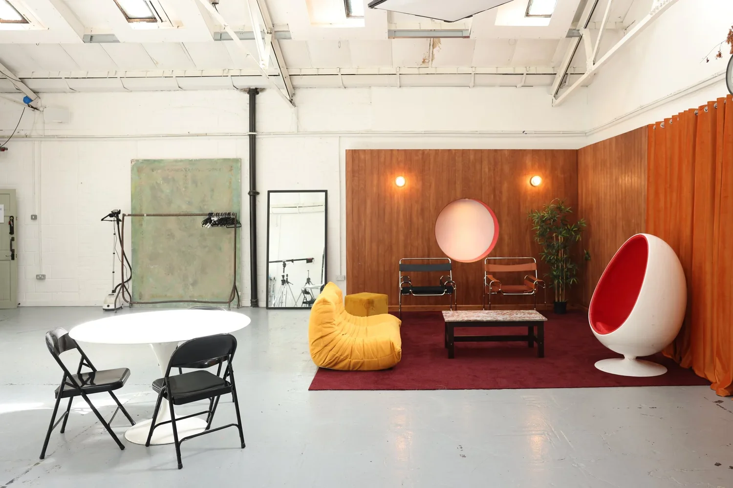 Interior of a studio or office space featuring a seating area with modern furniture, a yellow beanbag, chairs, a coffee table, decorative round light fixtures, a red egg-shaped chair, and a wooden wall with orange curtains.