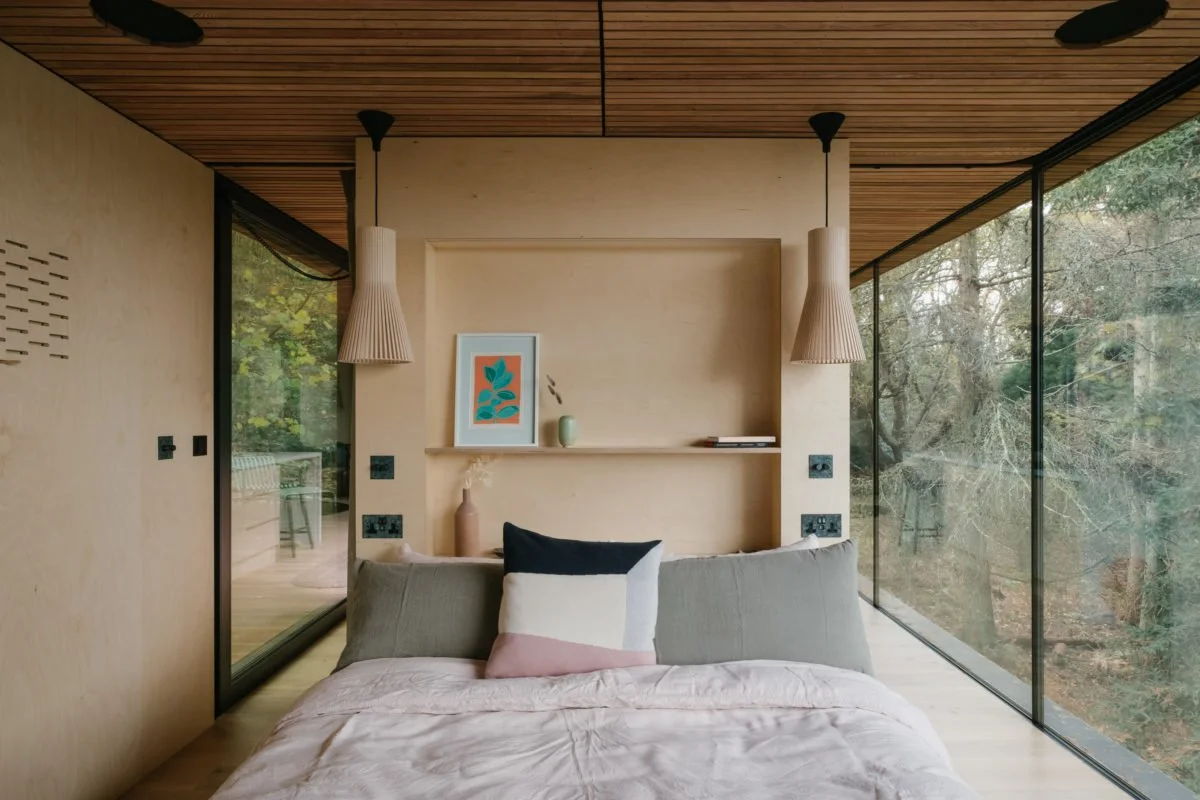 Bedroom with large glass windows showing trees outside, minimalistic decor, wooden ceiling, hanging pendant lights, bed with pillows, and wall shelf with framed art and vases.