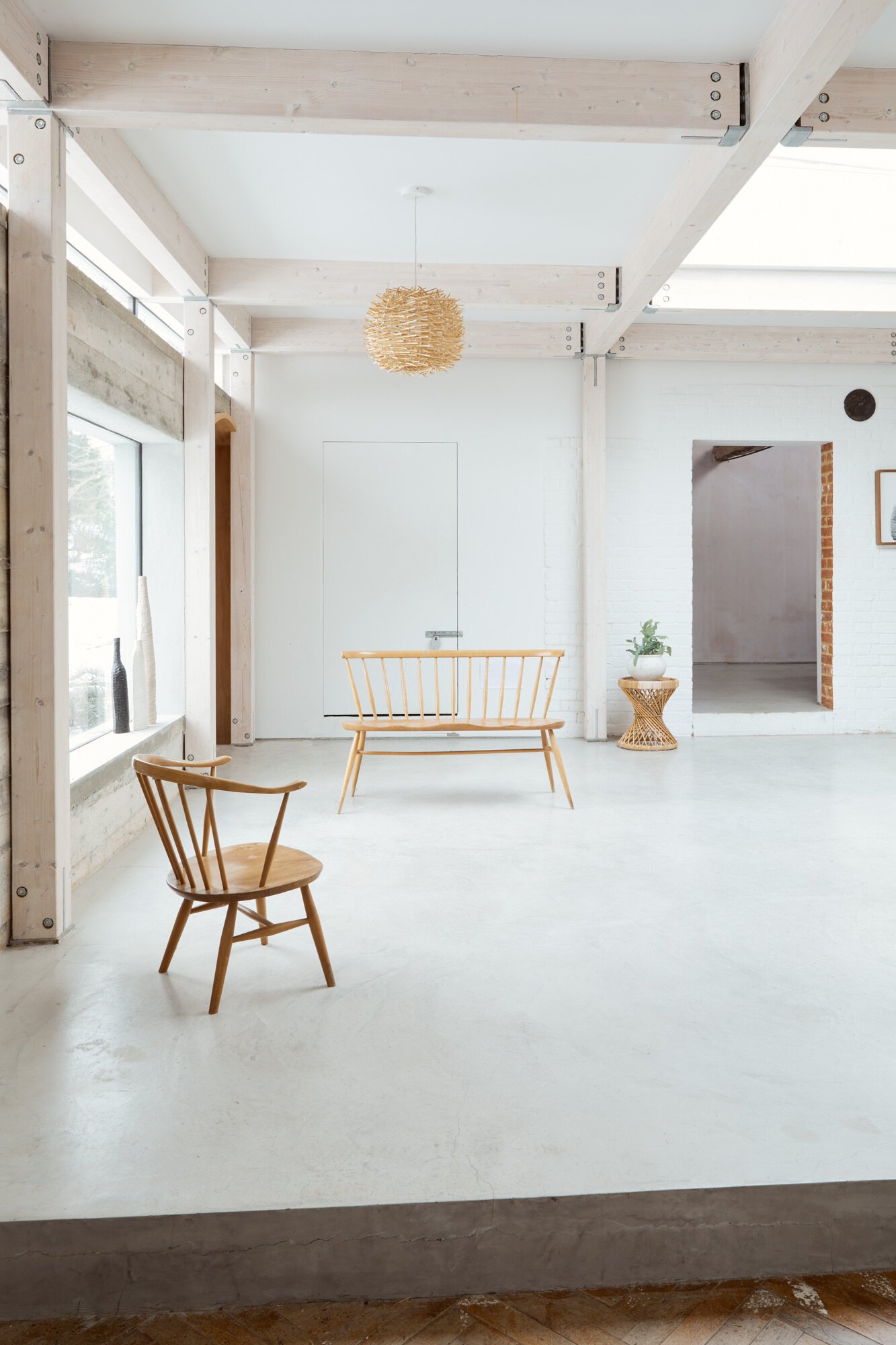 Minimalist interior of a bright room with white walls and light wooden beams. Two wooden chairs are placed on a white floor, with a window on the left and a decorative woven pendant lamp hanging from the ceiling. A small potted plant sits on a wicker