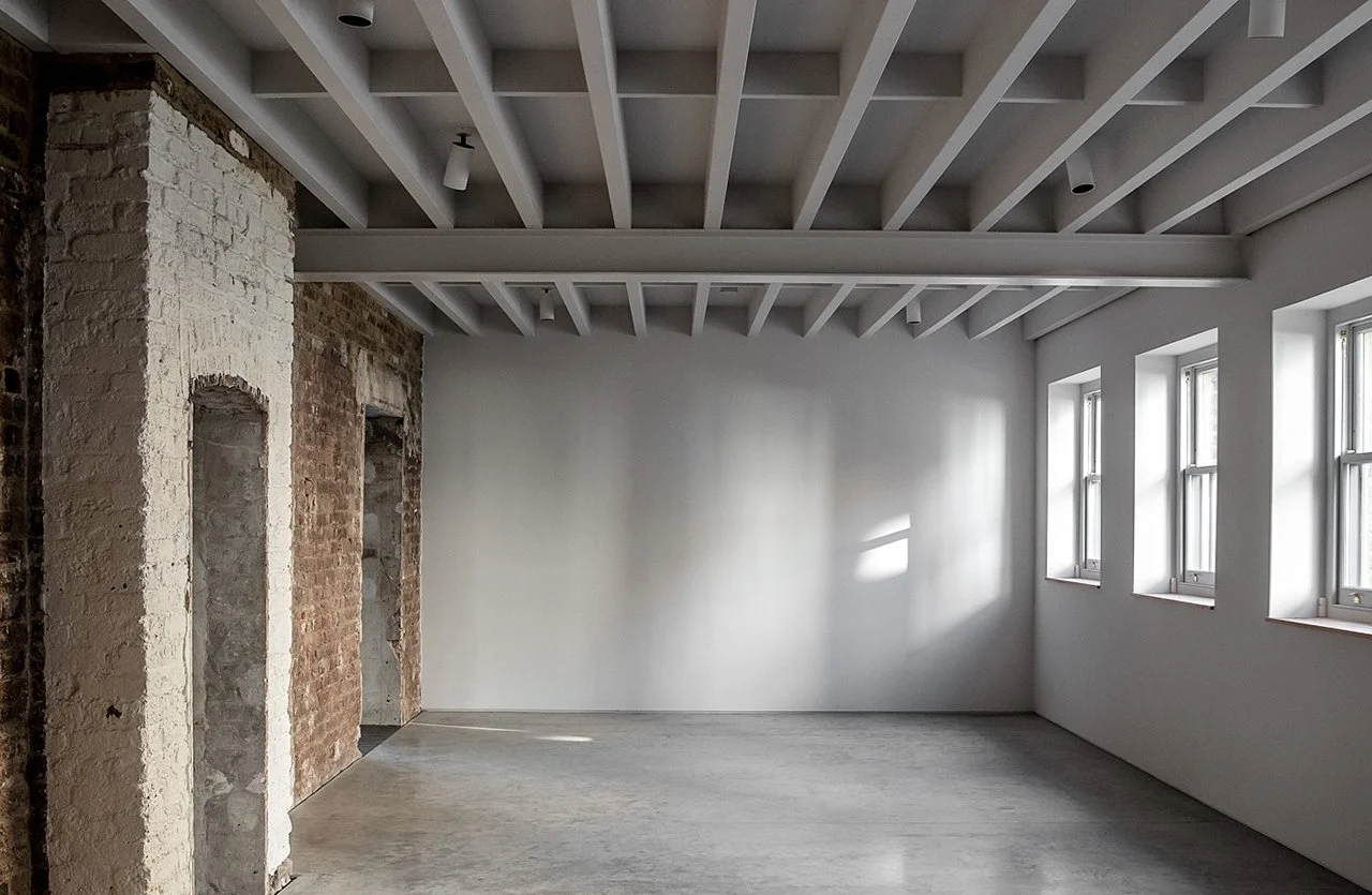 An empty room with white walls and a concrete floor, featuring exposed brick pillars and large windows allowing natural light to enter.