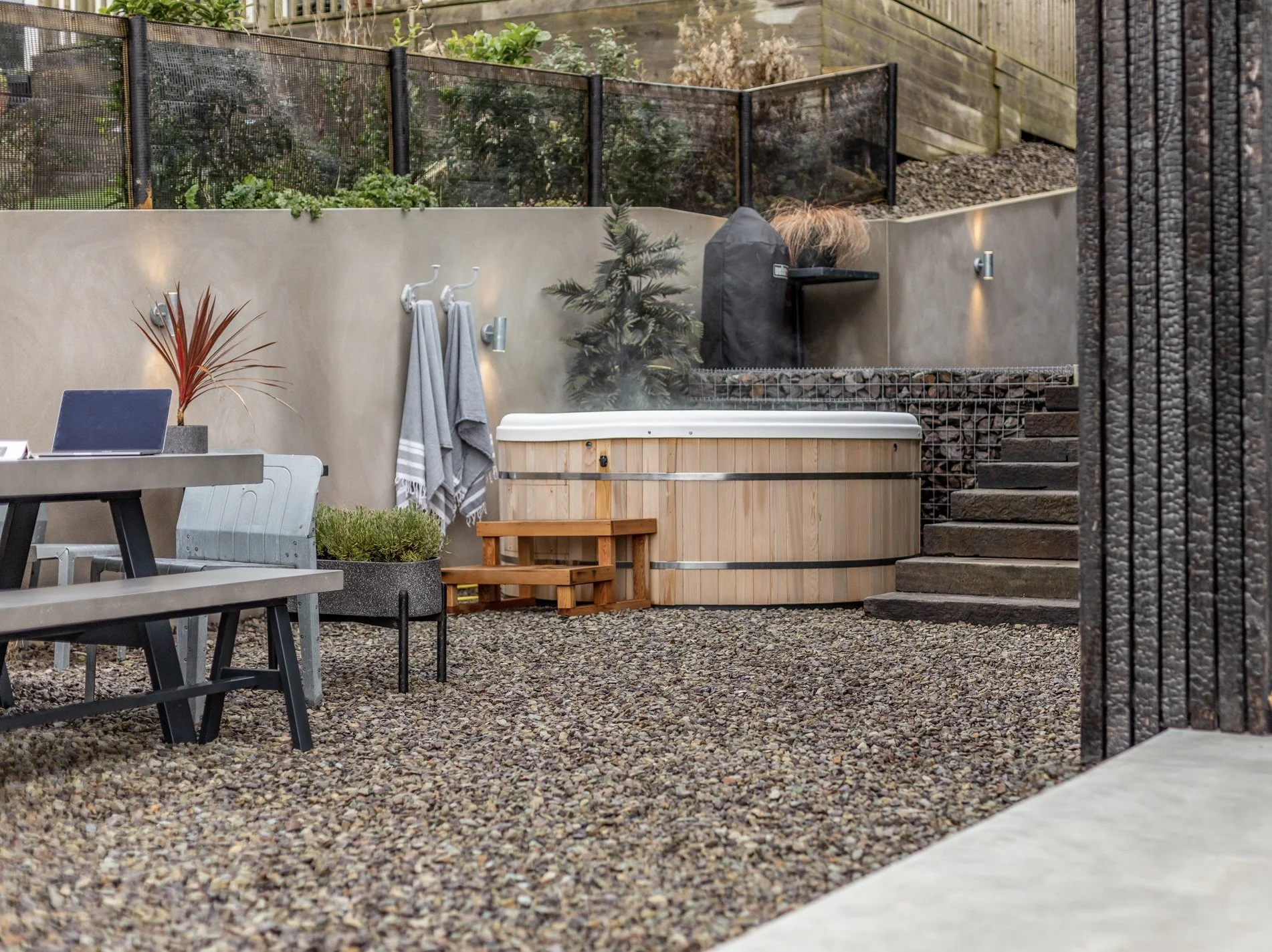 Outdoor backyard with a hot tub, a dining table with chairs, towels hanging on hooks, potted plants, a wood pellet grill, stone steps, and gravel ground.