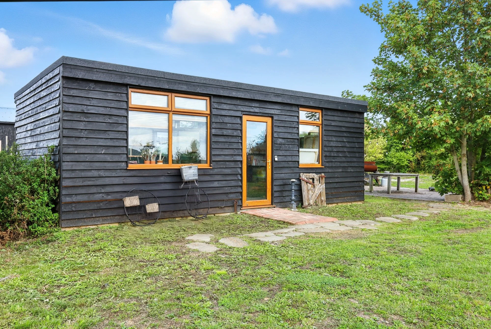 A small black wooden house with large windows and a wooden door, surrounded by green grass, trees, and a partly cloudy sky.