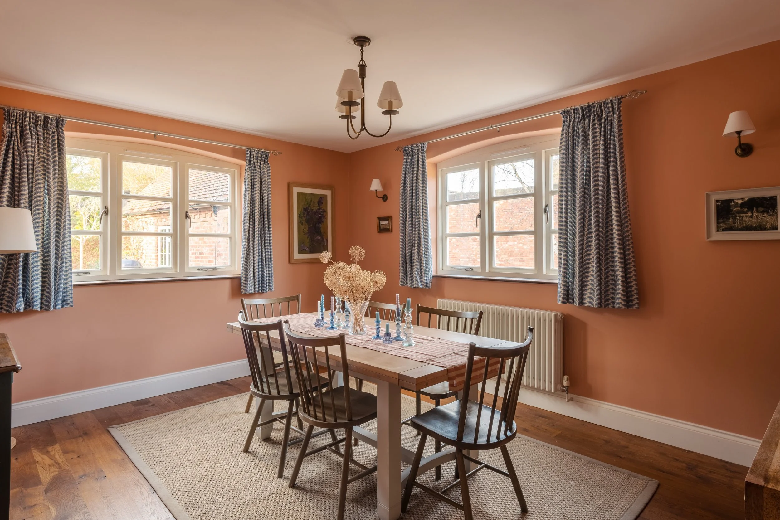 A cozy dining room with peach-colored walls, wooden flooring, four windows with patterned curtains, a dining table with six chairs, a vase with dried flowers, and framed artwork on the walls.