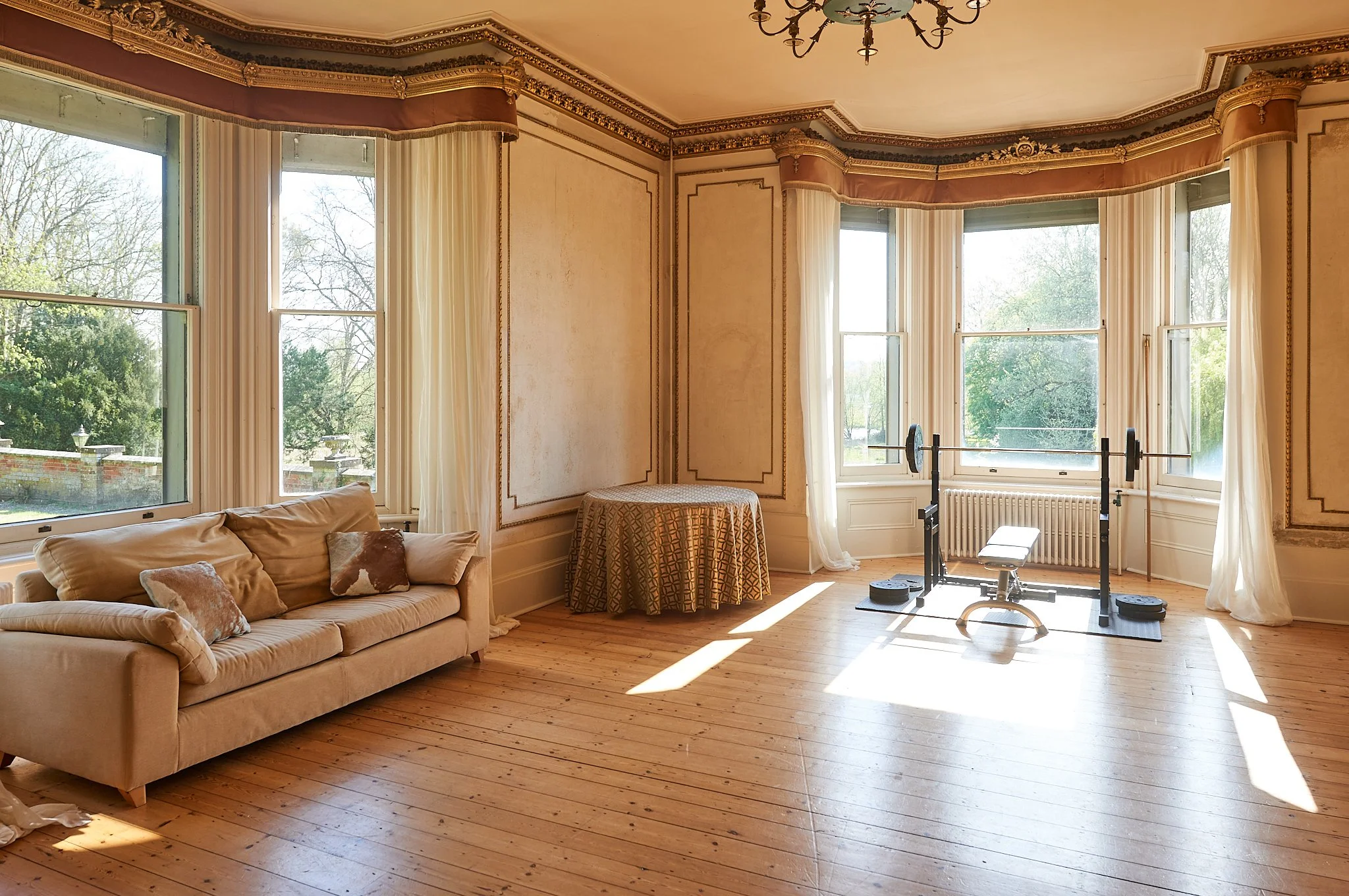 Living room with large bay windows, hardwood floors, a beige sofa with pillows, a small round table with a patterned cloth, and a weightlifting bench with weights.