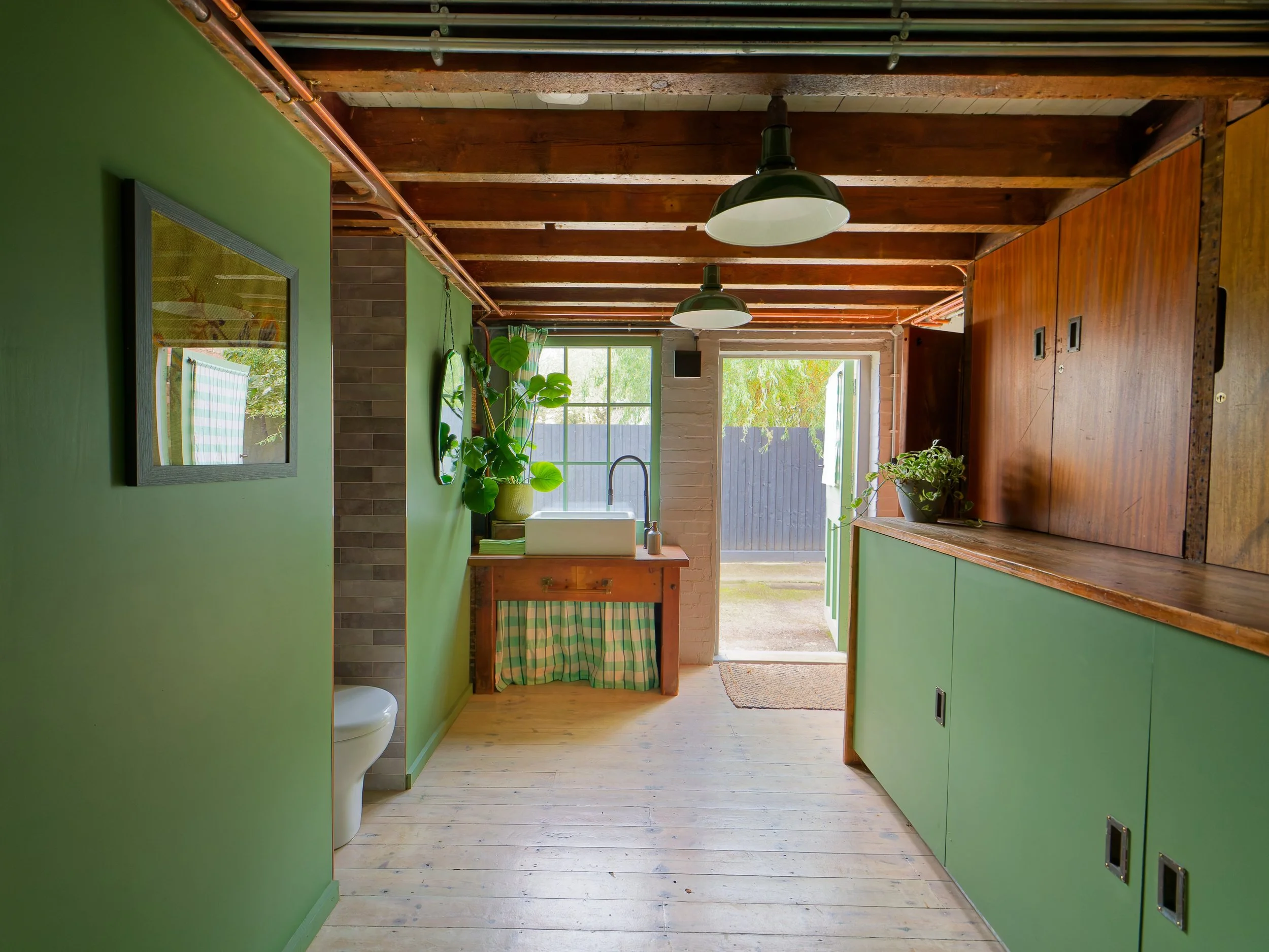 Interior view of a rustic kitchen with green walls, wooden ceiling beams, and sunlight streaming in through an open door. There are plants and a sink near the door, with kitchen cabinets on the right side.