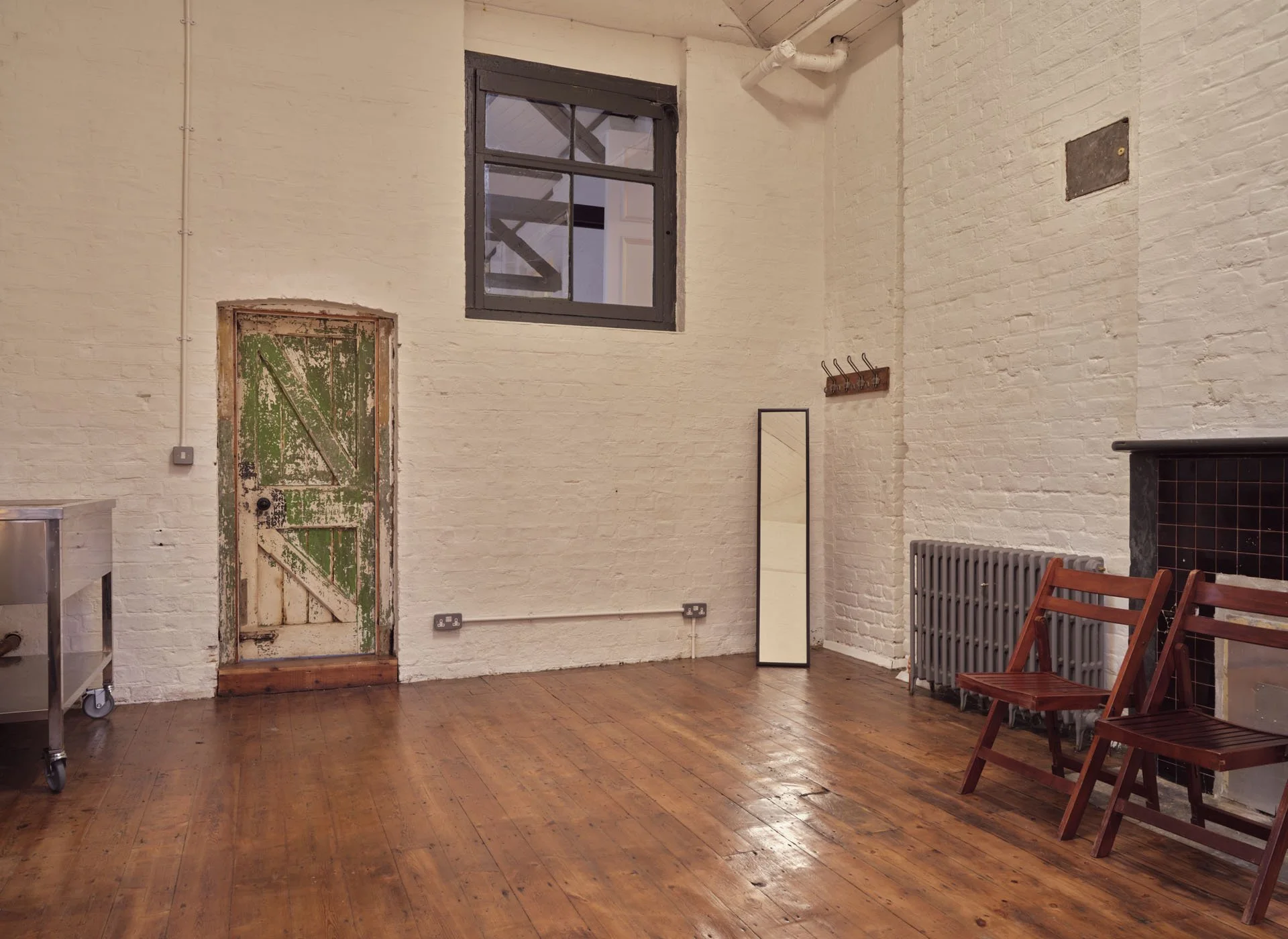 A room with white brick walls, wooden floor, a small distressed green door, a window, a mirror, wooden chairs, and a radiator.