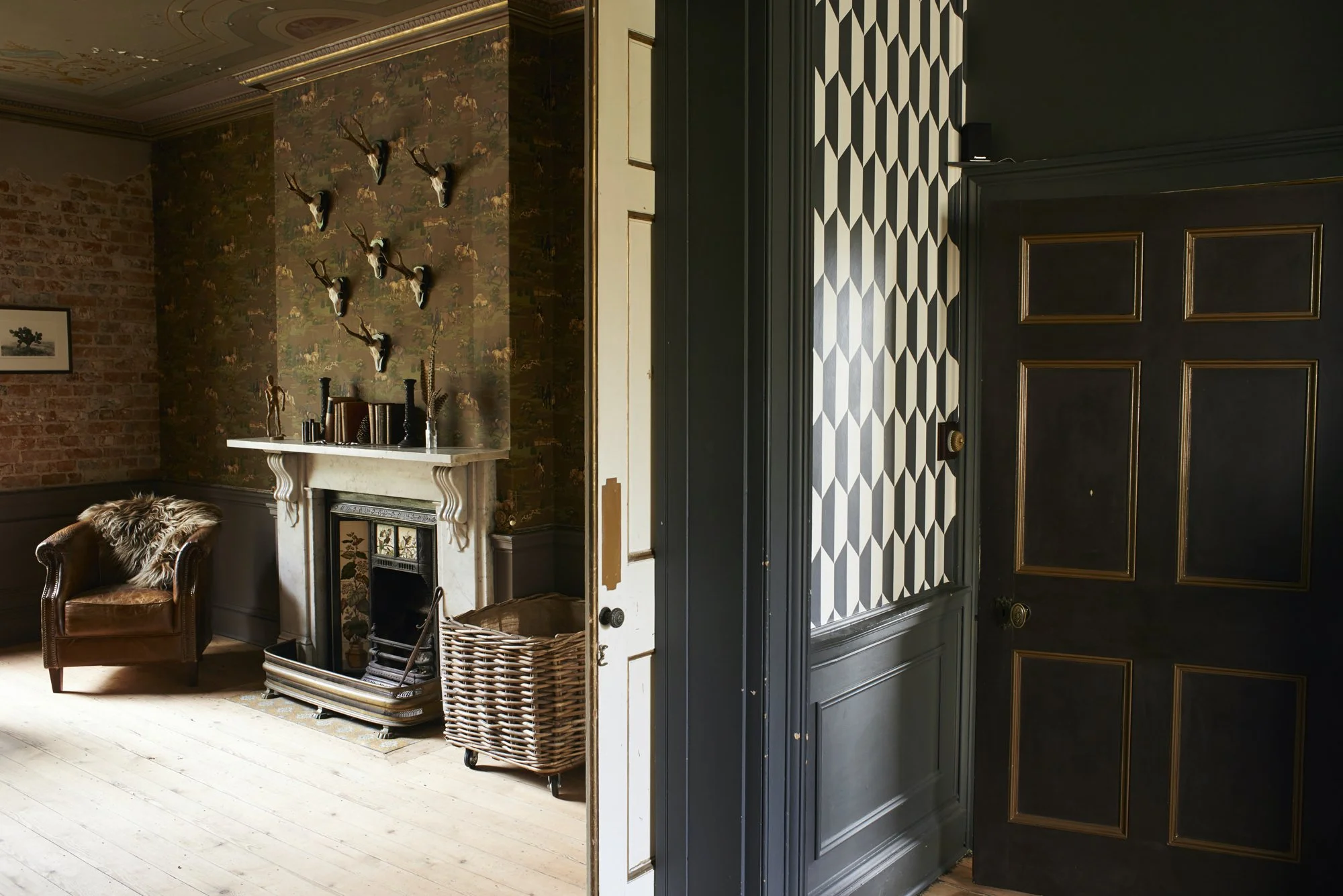 Interior of a stylish room with a brick and wallpapered wall, a fireplace with decorative objects, a brown leather armchair with a fur throw, and a basket beside the fireplace. To the right, part of a black paneled door and a patterned wall with geom