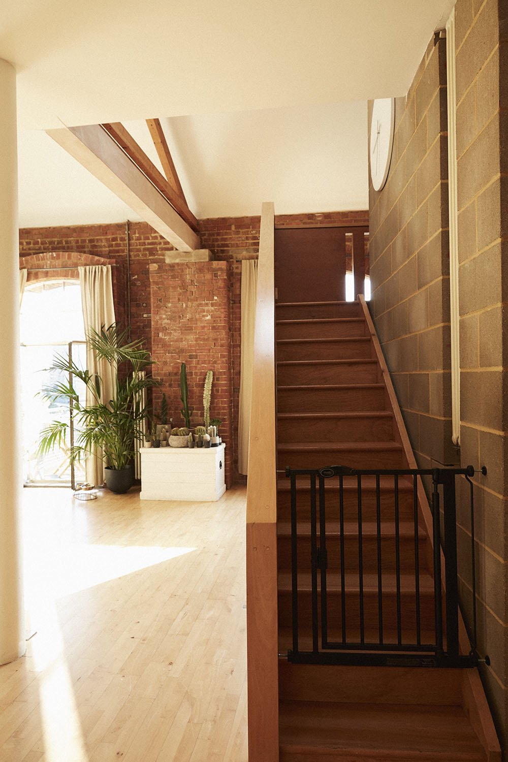 Interior of a house with wooden stairs, brick wall, large window with curtains, a green potted plant, and a white chest decorated with cacti.