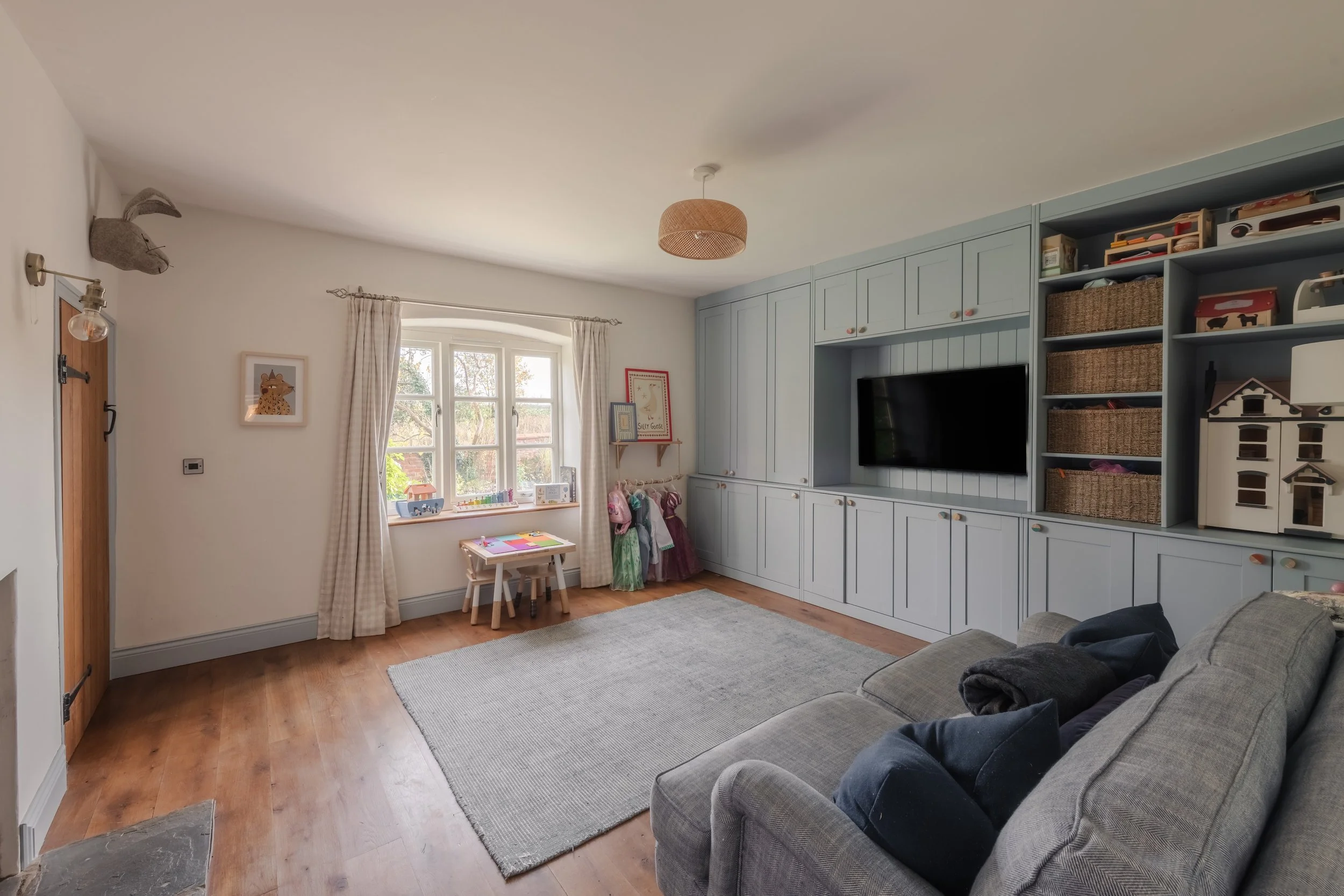 Living room with a gray sofa, built-in light blue cabinets, a wall-mounted TV, a window with curtains, a small table with children's books, toys, and dresses, and wall decorations.