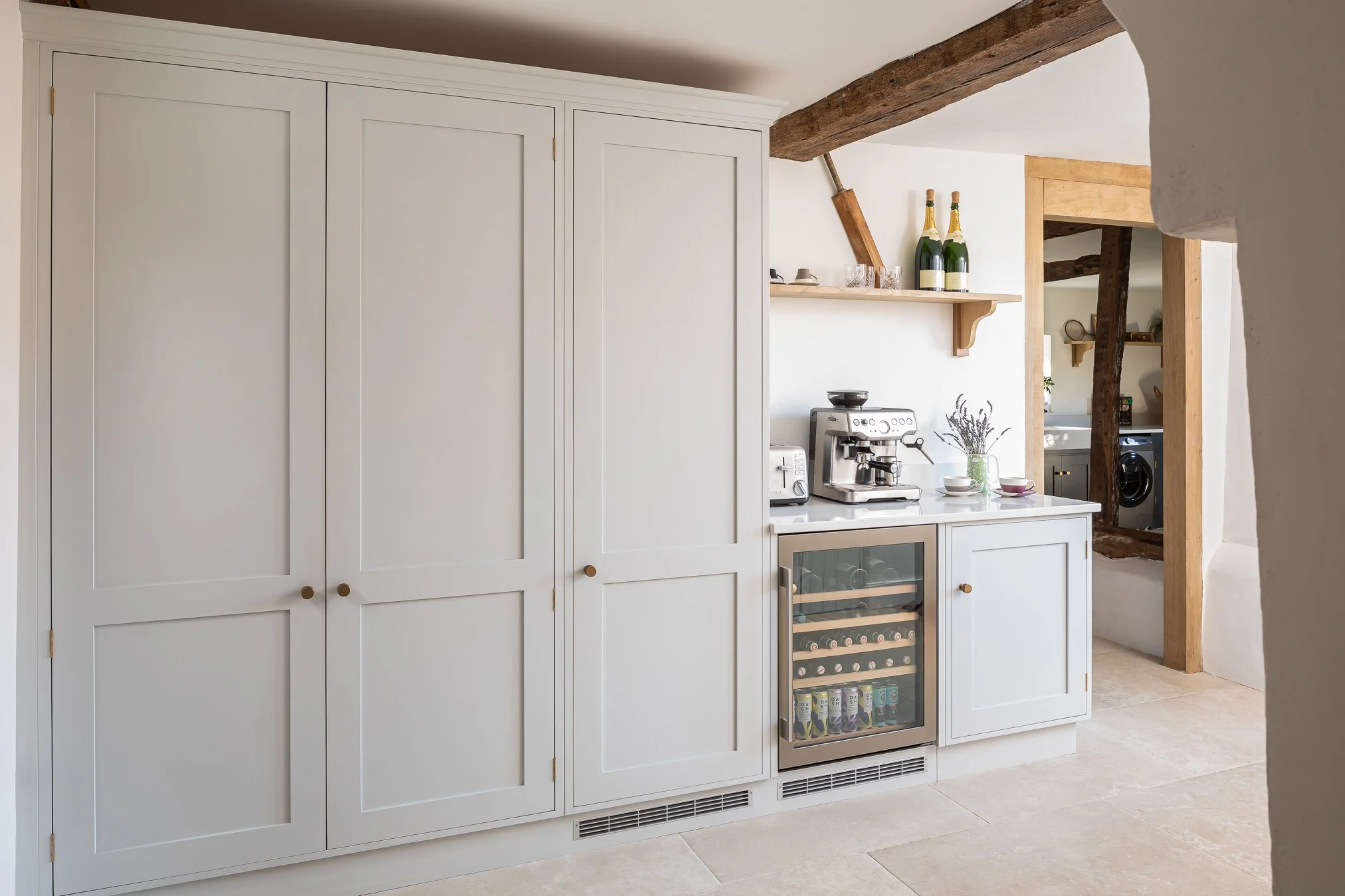 A kitchen with white cabinets, a small wine fridge, a coffee machine, and two bottles of champagne on a shelf, in a room with exposed wooden beams and white walls.