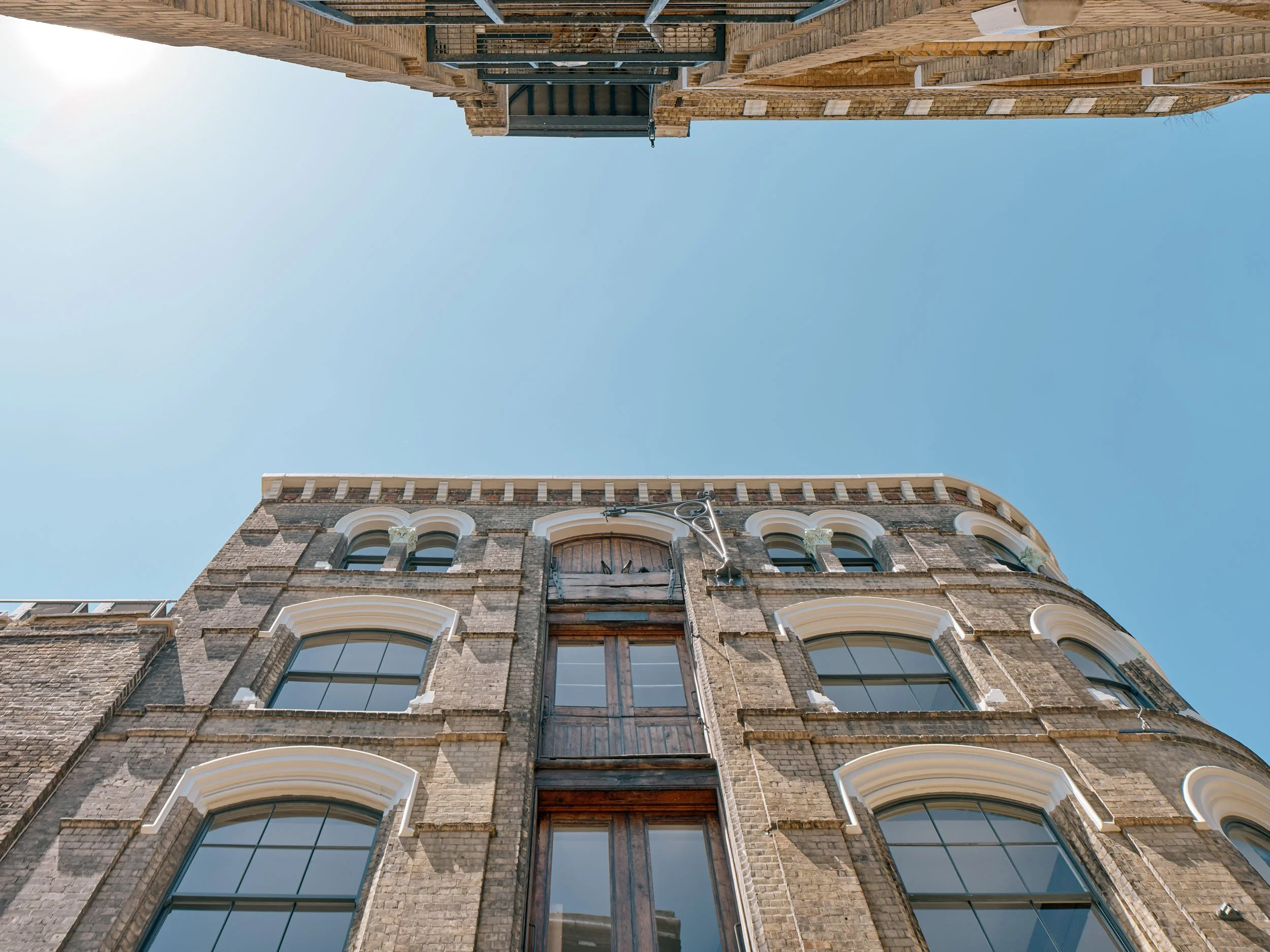 Looking up at the facade of old brick buildings with large arched windows and a clear blue sky above.