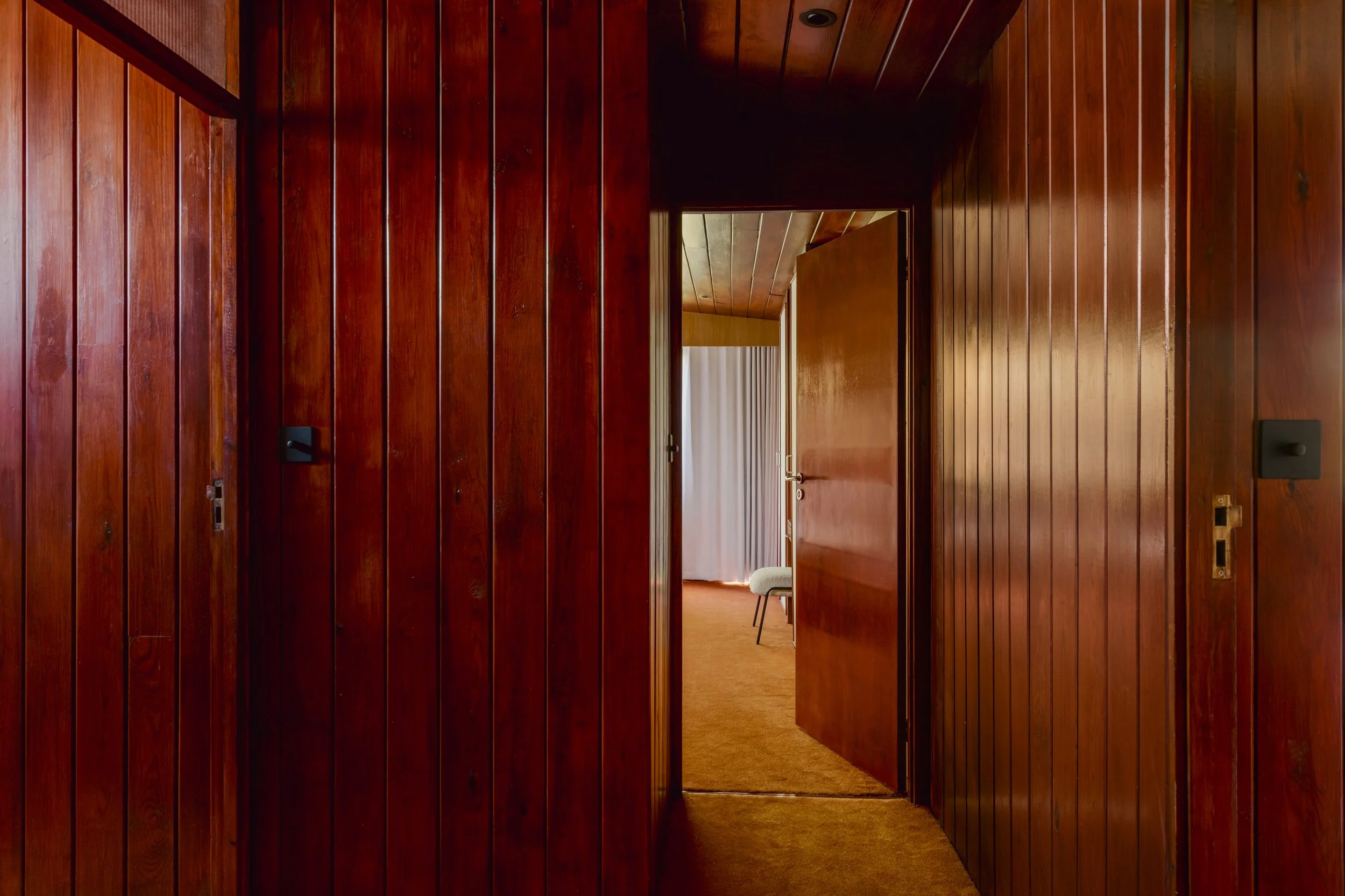 Wood-paneled hallway with two open doors leading into a room with a white curtain and chair.