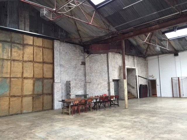 An empty industrial indoor space with a high ceiling, exposed beams, and a concrete floor. There is a long table with chairs along one wall, and a staircase leading up to another level.
