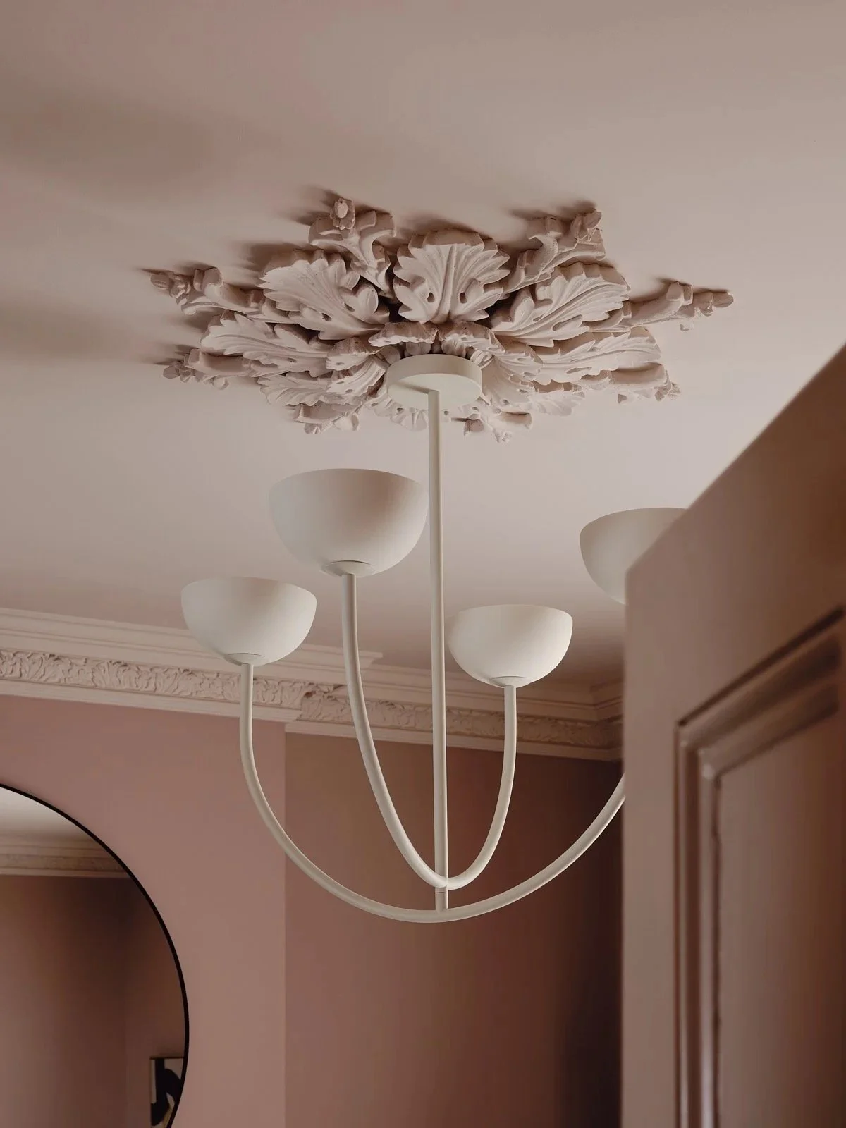 A ceiling light fixture with five white, bowl-shaped lamps. The fixture hangs from a decorative ceiling medallion in an interior room with beige walls and crown molding.