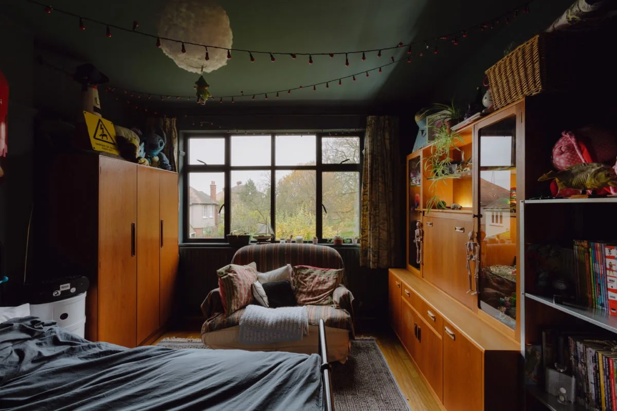 Cozy living room with a large window, a couch with pillows, wooden cabinets, and a bed in the foreground. Decor includes hanging string lights and plush toys on top of the cabinet.