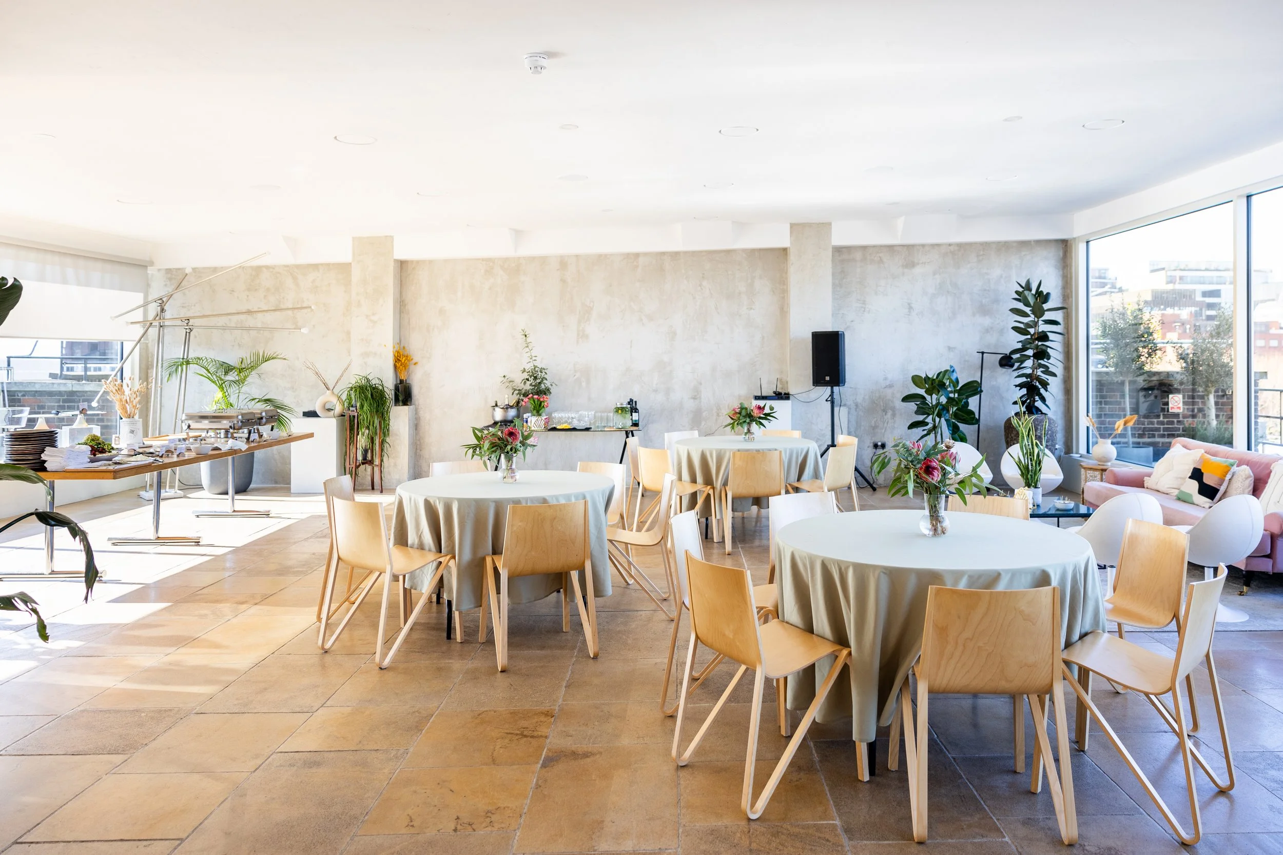 A bright, modern banquet hall with wooden tables and chairs, decorated with floral centerpieces, and large windows letting in natural light.