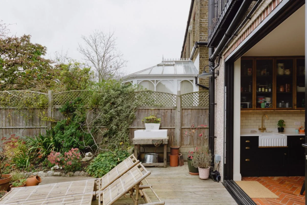 View of a backyard patio with outdoor furniture, potted plants, garden tools, and a view into a kitchen with black cabinets and a gold faucet.