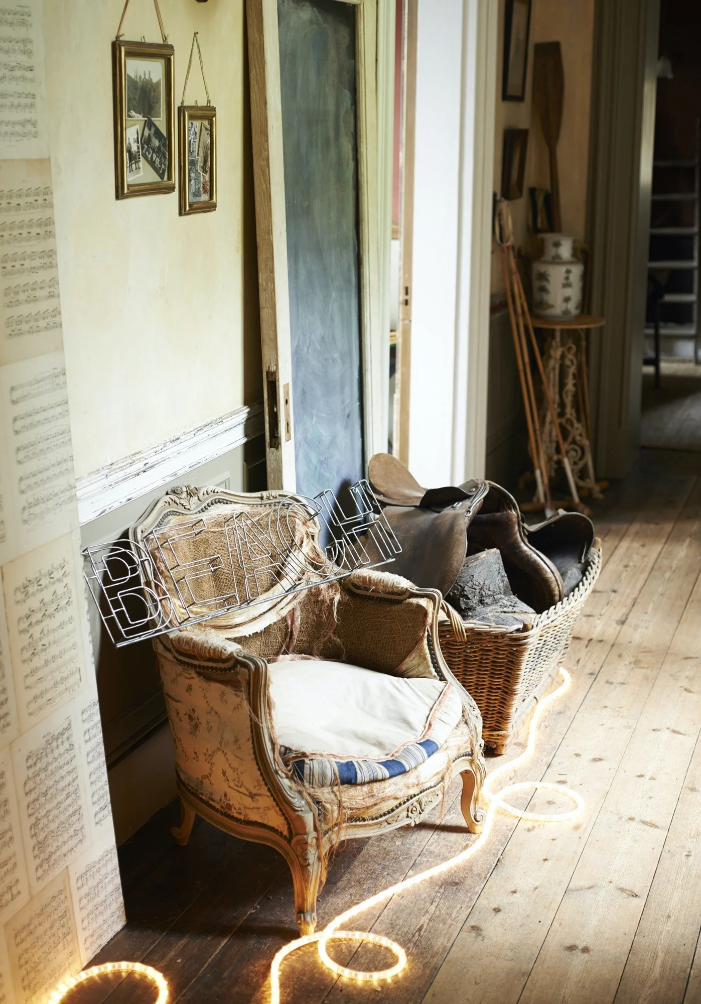 Vintage living room corner with ornate armchair and wicker basket filled with used cushions and a saddle, illuminated by string lights on the wooden floor, with framed photos hanging on a yellow wall and a chalkboard leaning against the wall.