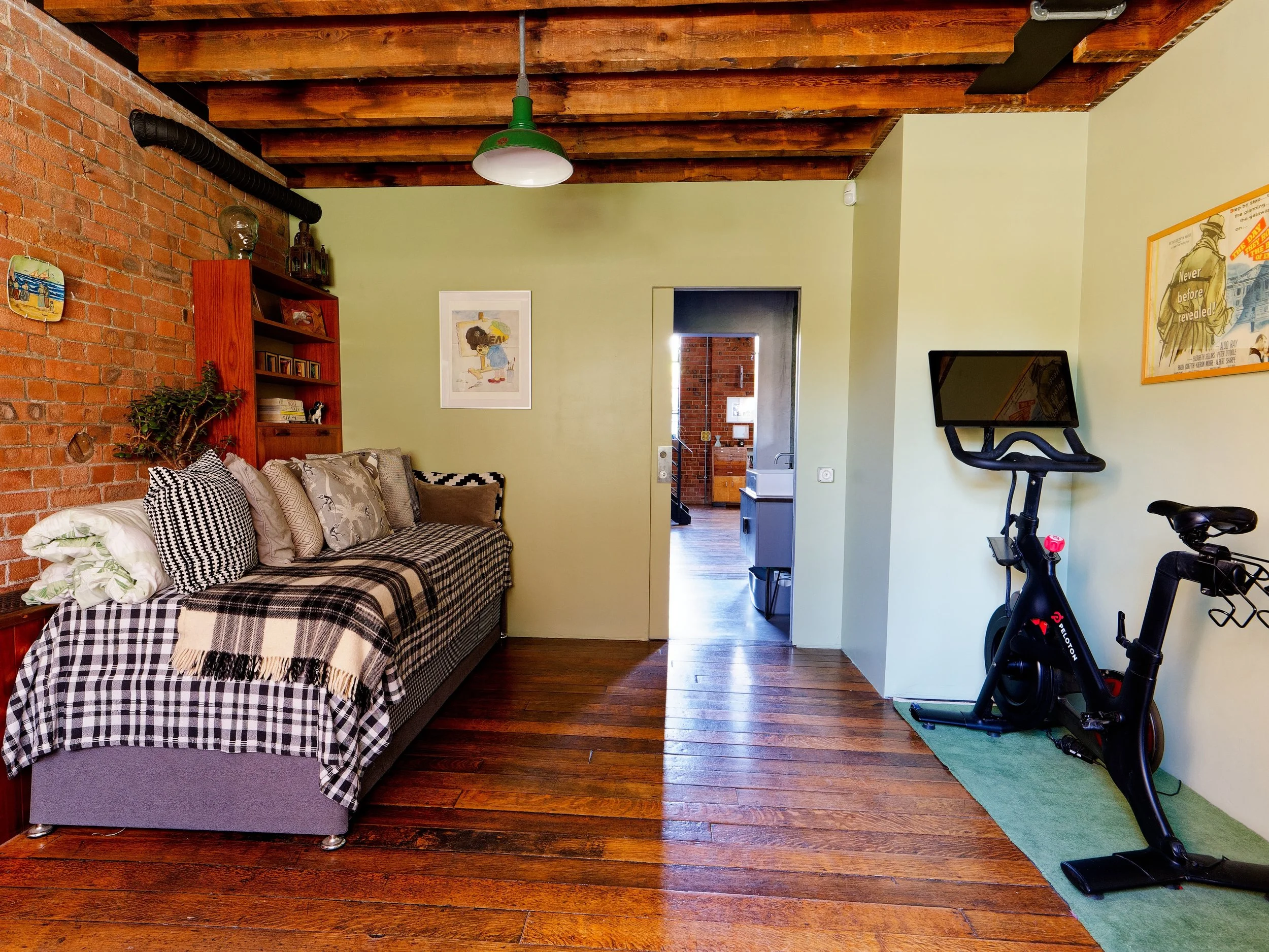 A living room with a brick wall, a green wall, a gray sofa with checkered pillows and a tartan blanket, a wooden shelf, framed artwork, a doorway revealing a kitchen, and an exercise bike on a green mat.