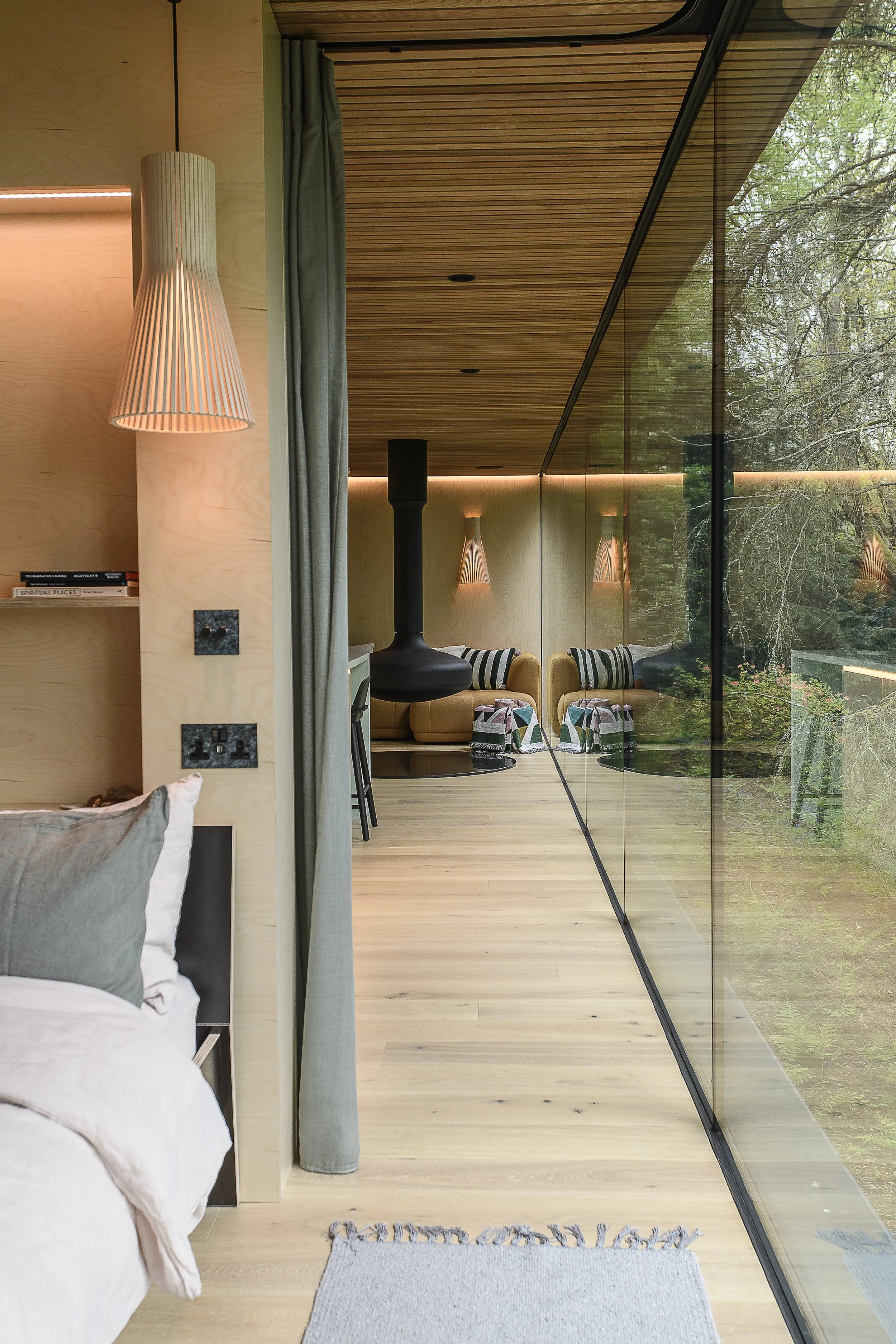Interior view of a modern bedroom with a large glass wall showing trees outside, a bed with pillows, a bedside lamp, a built-in bookshelf, and a living area with yellow sofas and striped pillows in the background.