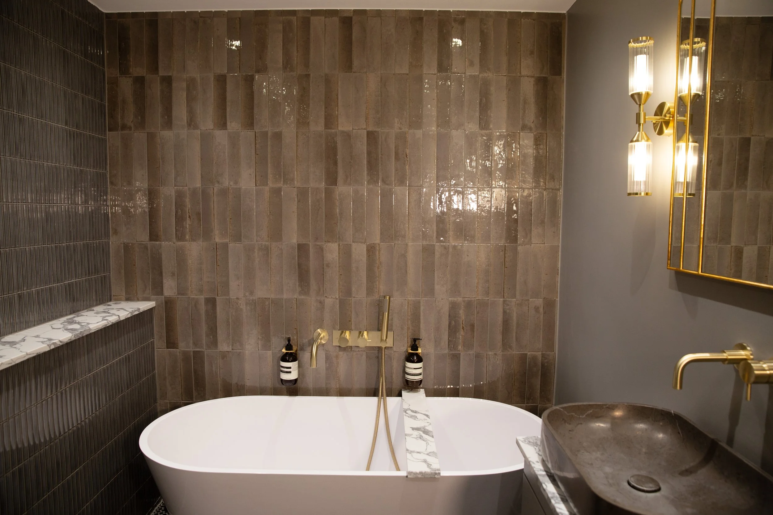 Modern bathroom with a freestanding bathtub, brown tiled wall, gold fixtures, marble shelf, and a sink with gold faucet.