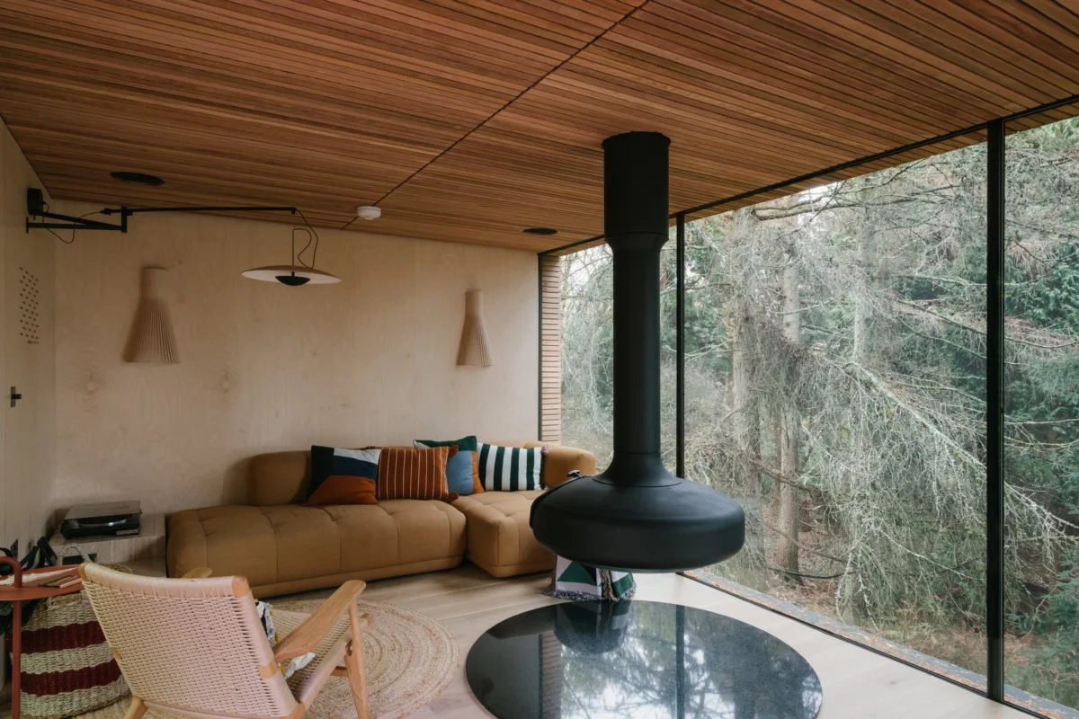 Interior living room with a beige sectional sofa, colorful pillows, wooden ceiling, large glass windows showing trees, and a black fireplace hanging from the ceiling.