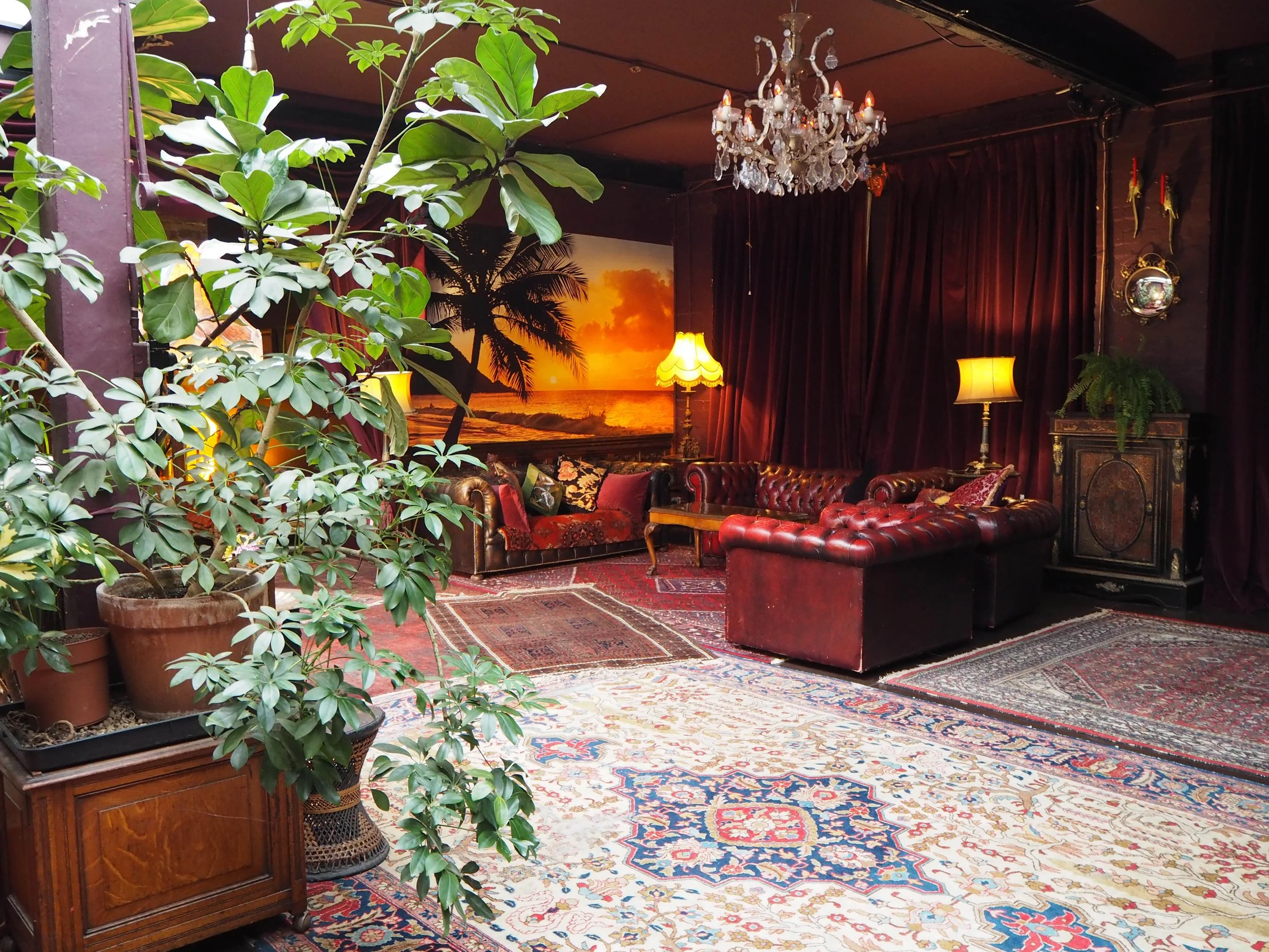Interior of a cozy living room with dark red curtains, vintage leather sofas, a chandelier, and potted plants, with a sunset beach scene painting in the background.