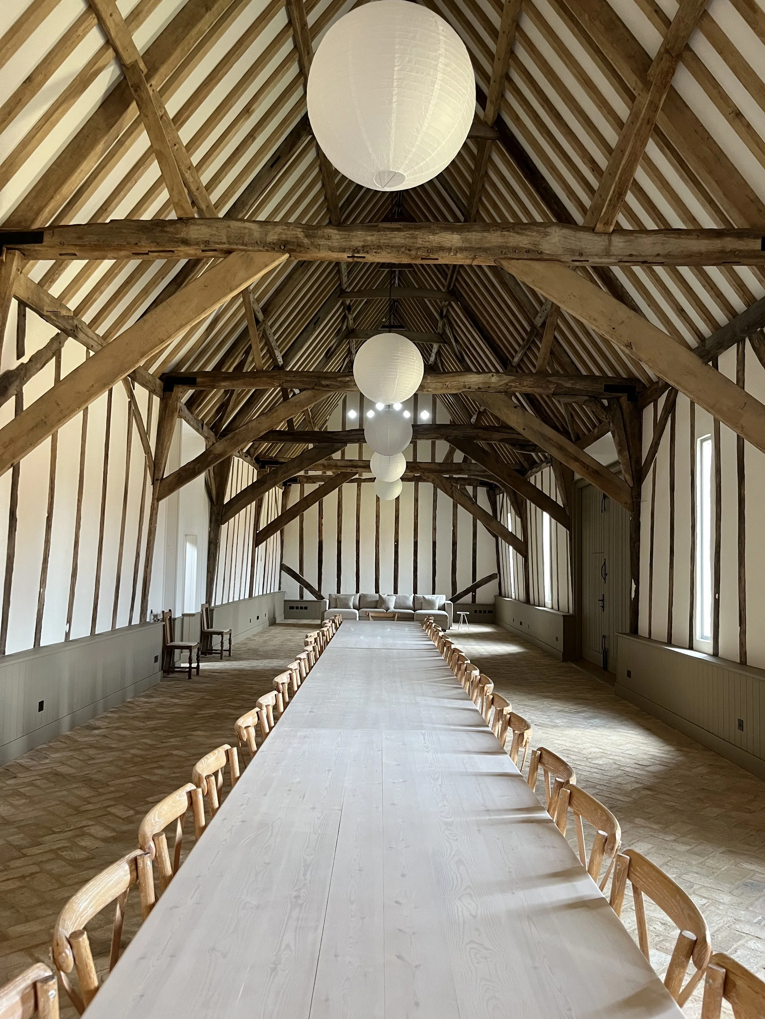 A long wooden table with chairs in a rustic, large attic or barn-like room with exposed wooden beams, paper lantern light fixtures, and a row of windows on the right wall.