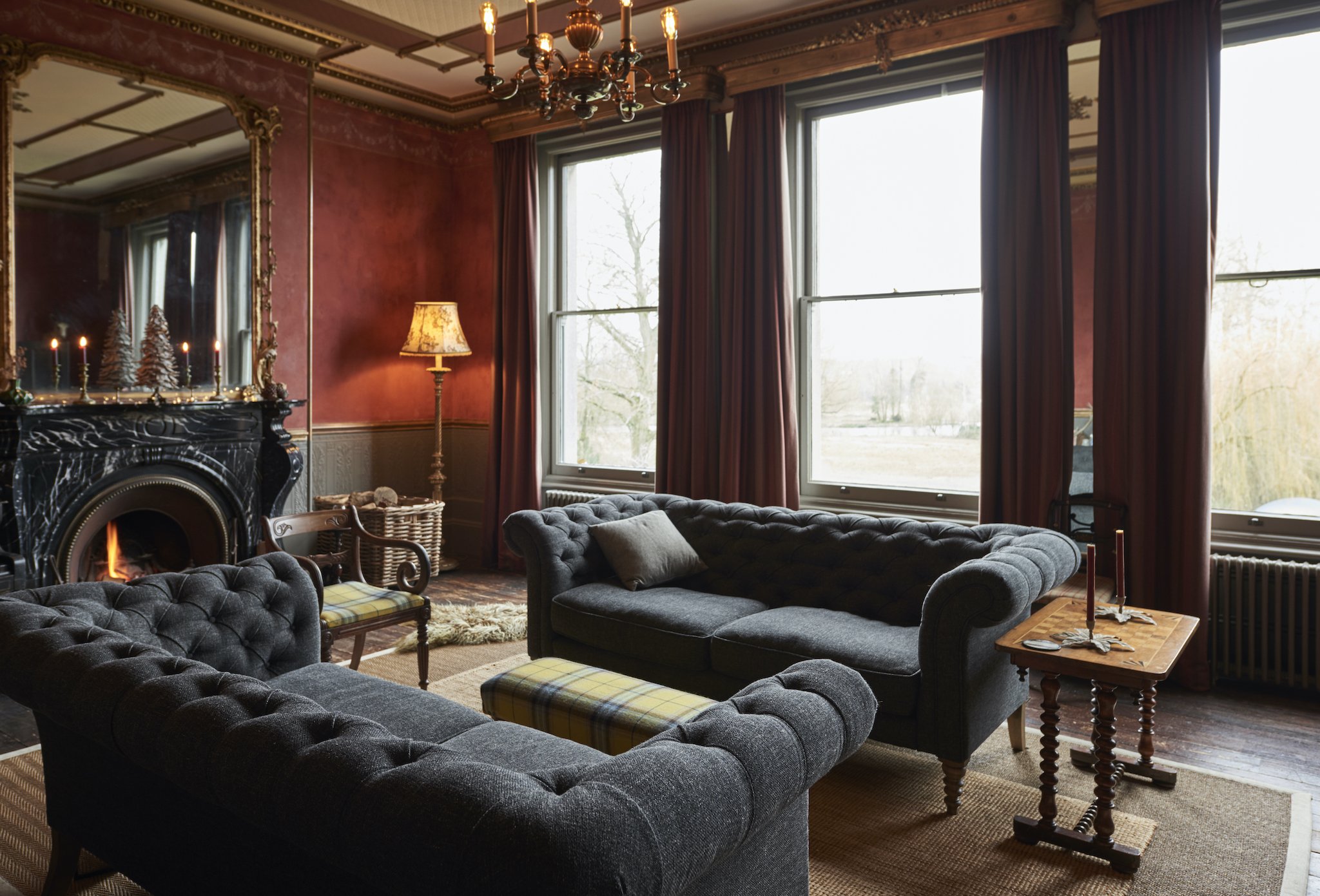 A cozy living room with dark gray tufted sofas, a wooden side table with candles, a standing lamp, a decorative mirror above a black marble fireplace, and large windows with dark red curtains, overlooking a landscape outside.