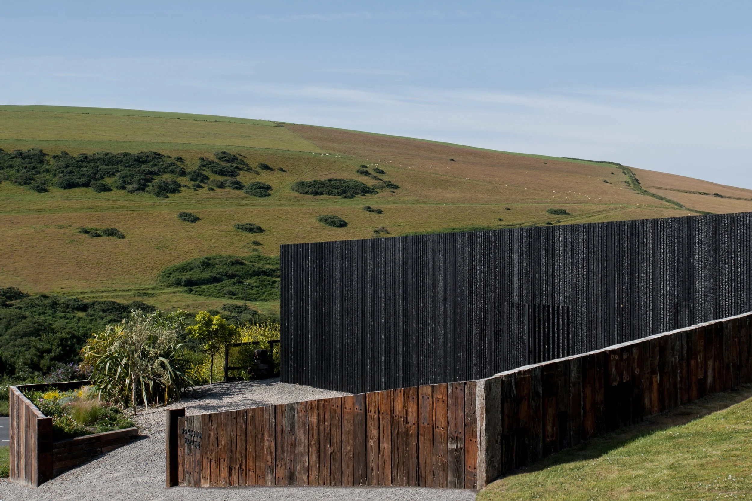 A modern outdoor structure with wooden and black metal fencing, set against a backdrop of rolling green hills and a clear sky.