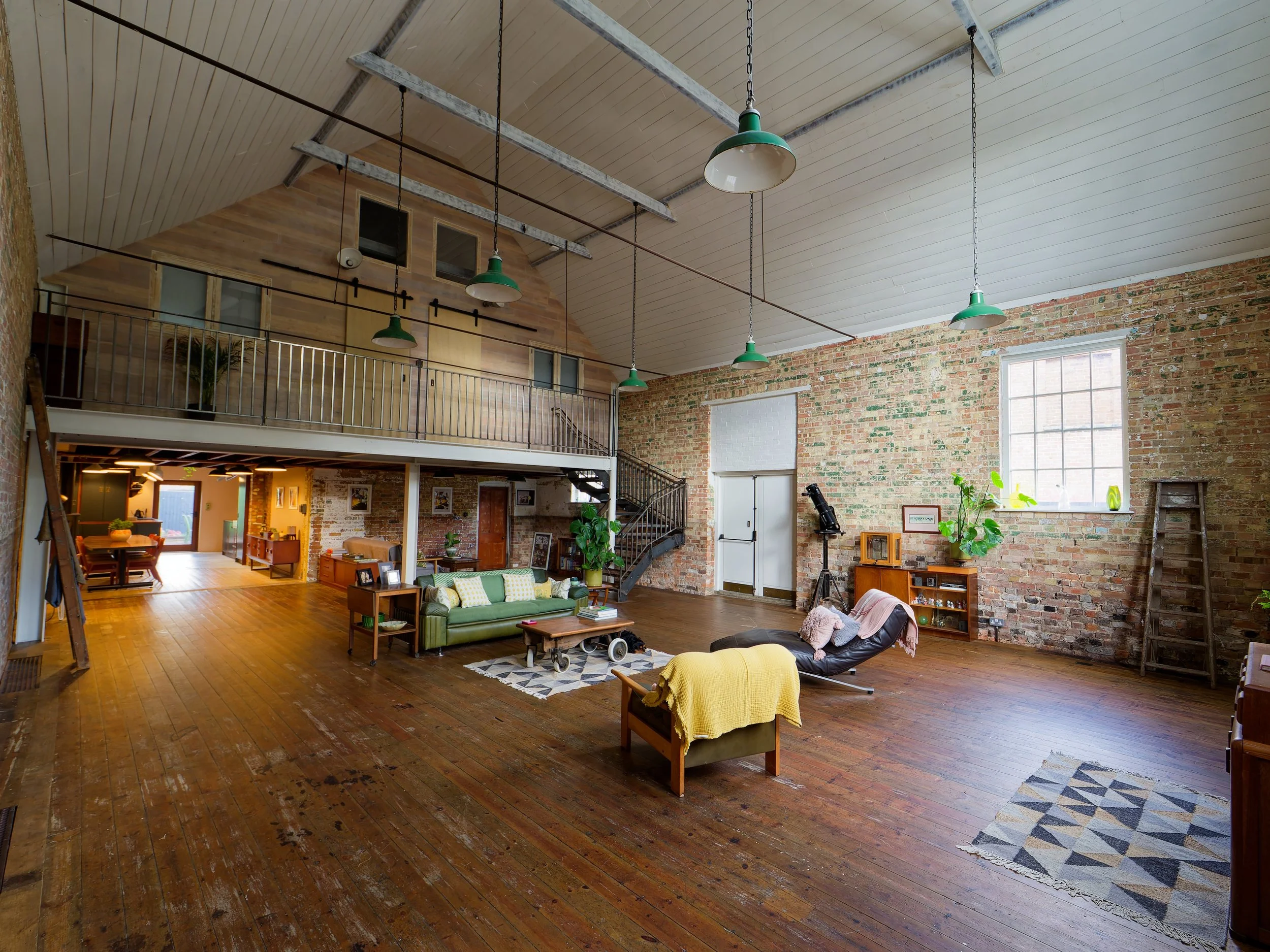 Spacious loft-style living room with exposed brick walls, wooden floors, and high ceilings. Furniture includes a green sofa, black lounge chair, and wooden coffee table. The room is decorated with potted plants, rugs, and vintage items. There are lar