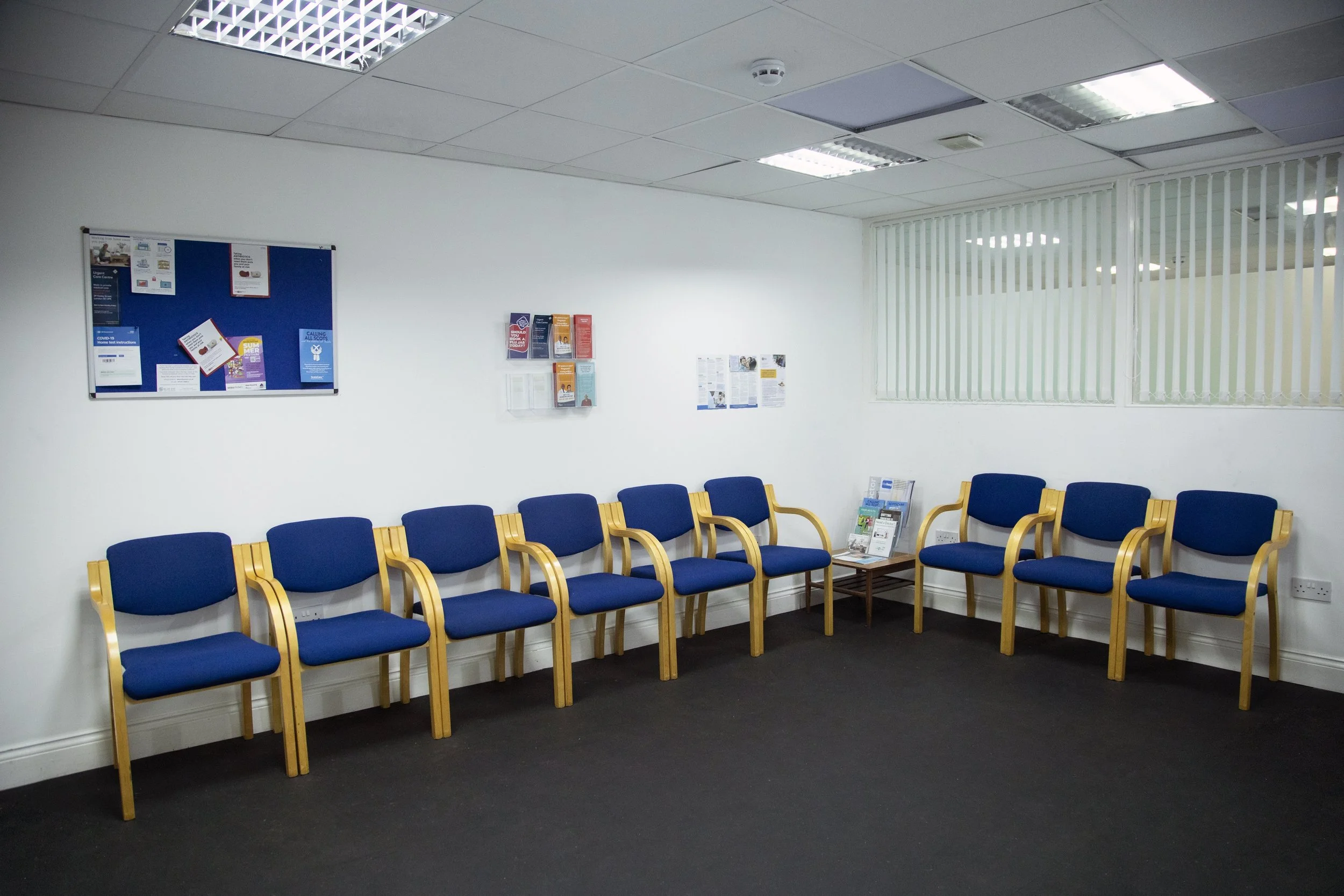 A waiting room with ten blue cushioned chairs with wooden armrests, arranged in two rows along the wall, a small table with magazines, white walls, bulletin boards on the wall, and a window with vertical blinds.