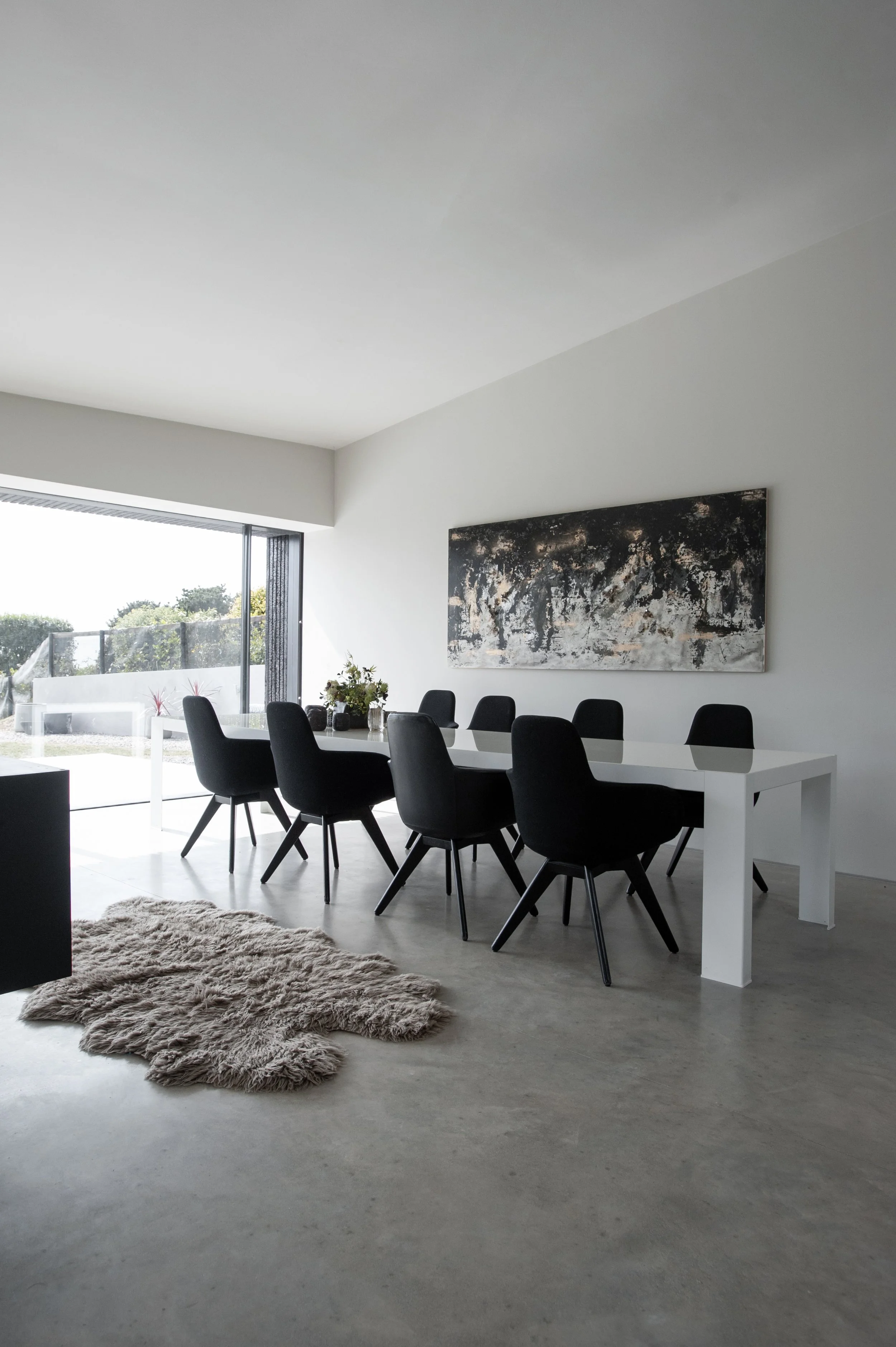 Modern dining room with a white table, eight black chairs, a shaggy beige rug, a large abstract painting on the wall, and a sliding glass door leading outside.