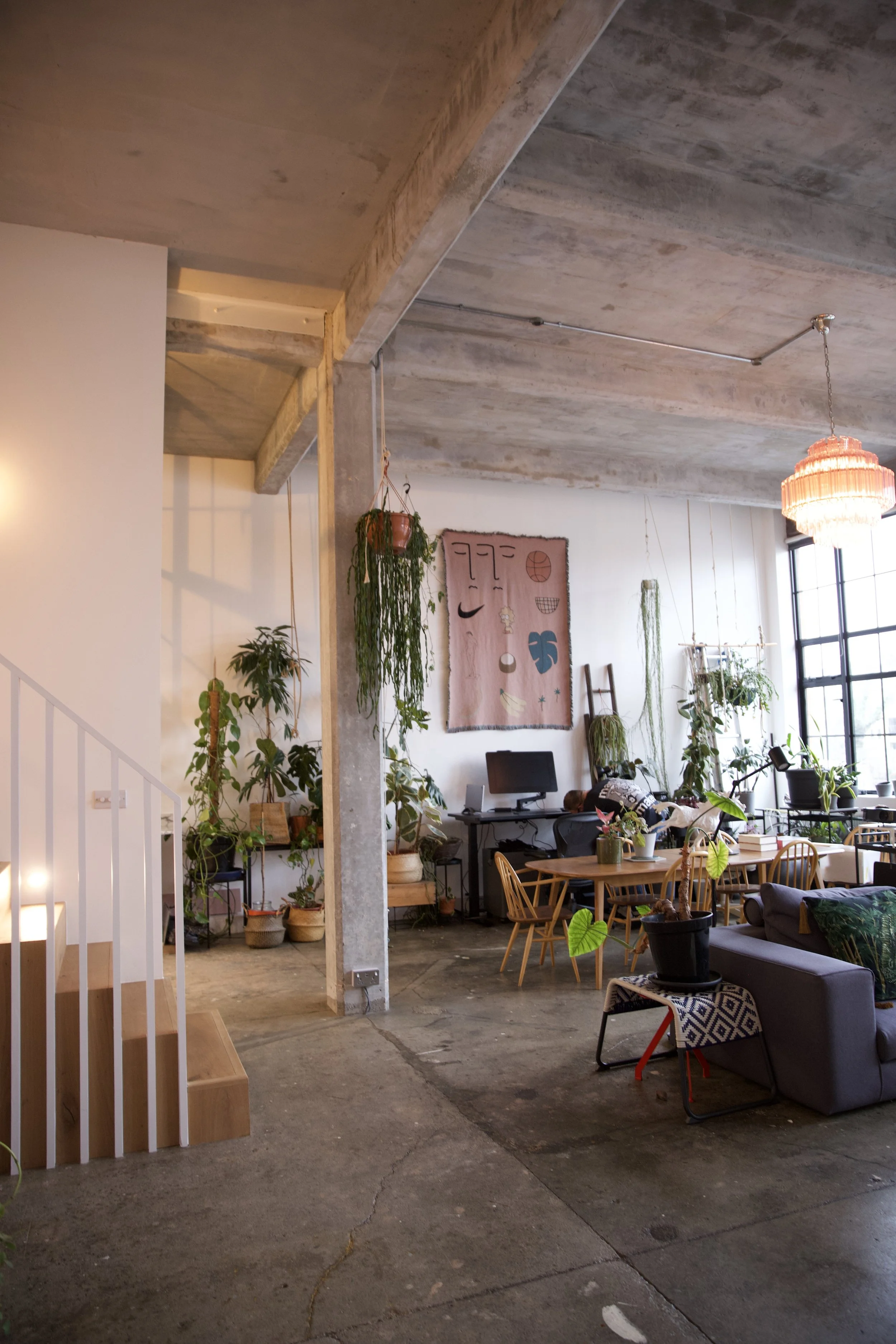 A spacious and modern loft-style living area with industrial concrete floors and ceiling, decorated with numerous indoor plants, a wall hanging art piece, a computer desk, and a dining area with wooden chairs and a table, illuminated by a large windo