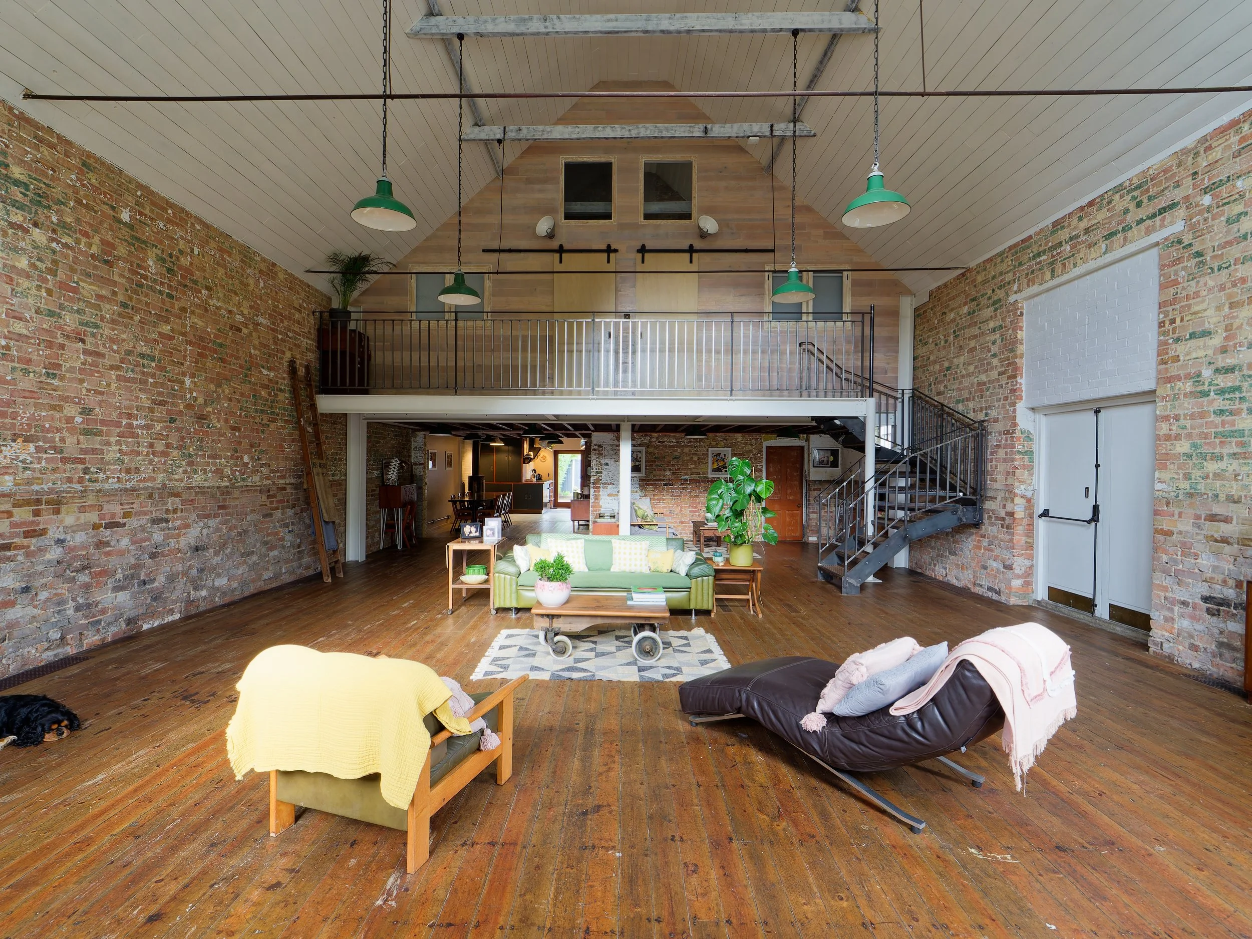 Spacious loft-style living room with exposed brick walls, wooden floors, and an open mezzanine level. Furniture includes a green sofa, a wooden armchair with a yellow throw, and a brown reclining chair with pillows and a pink throw. There are multipl