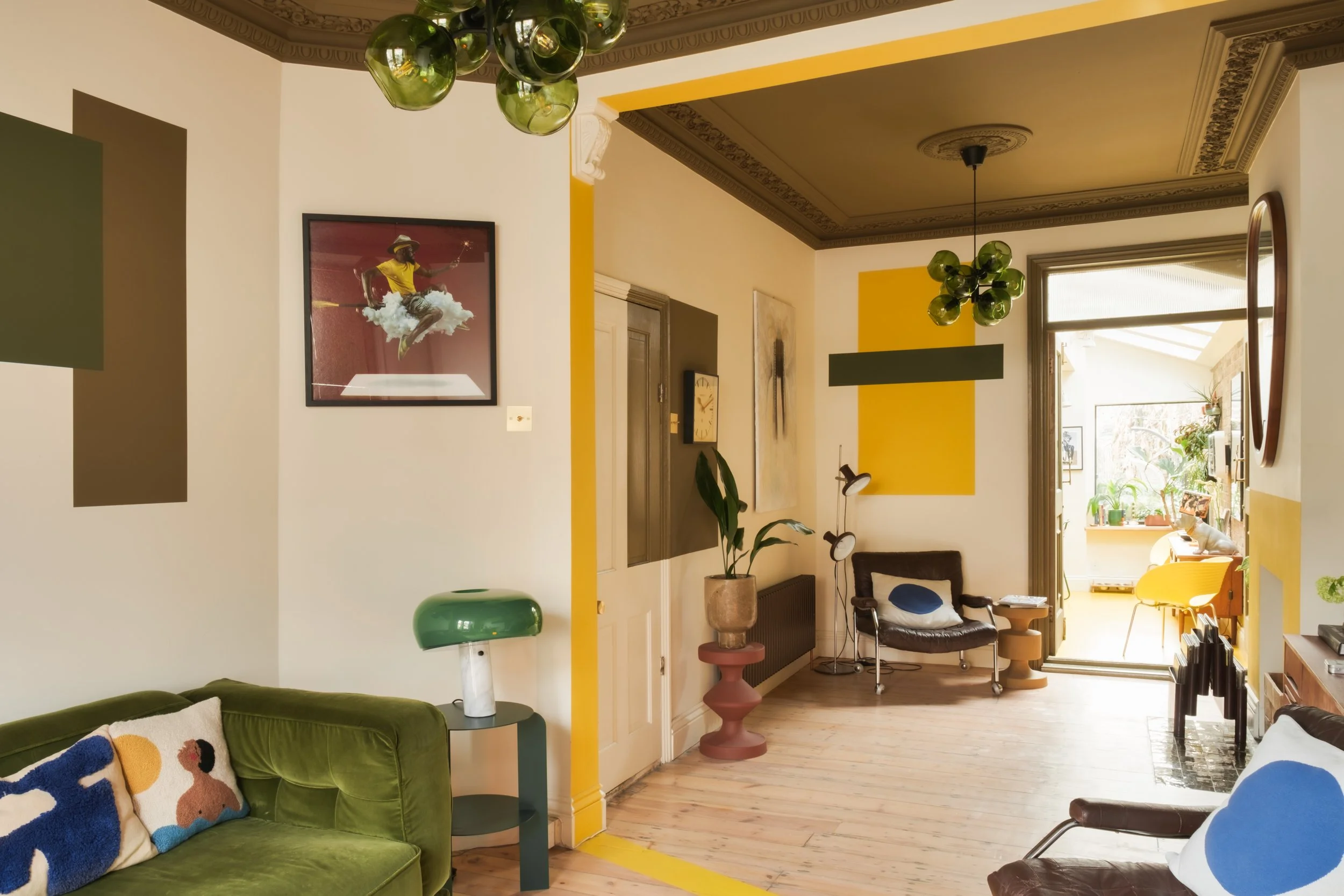 Living room with green sofa, framed art on the wall, potted plant, lounge chairs, and a sunlit balcony with plants.