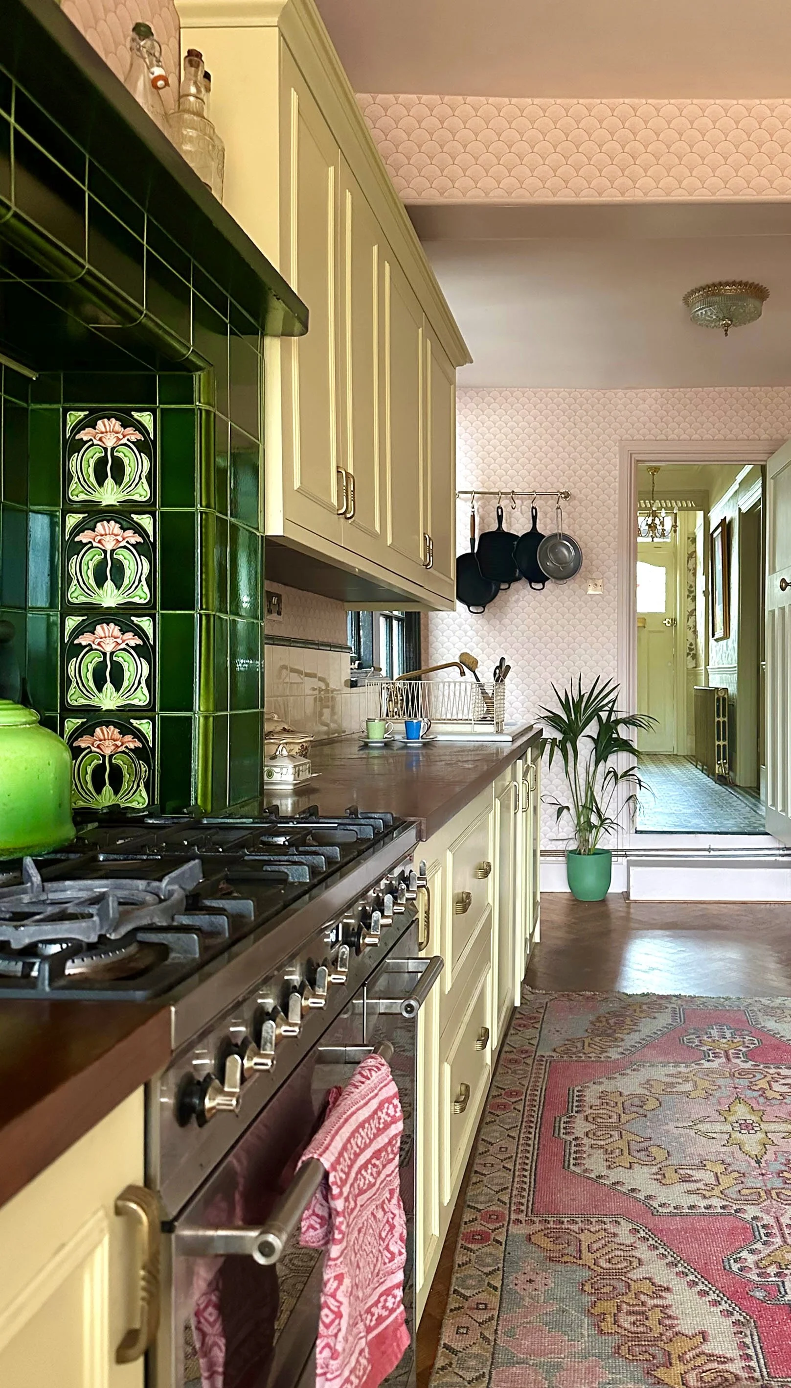 A cozy vintage kitchen with cream cabinets, a dark wood countertop, a green tiled stove with floral motifs, a pink patterned wallpaper, and a kitchen rug. There are hanging pots and a potted plant near the doorway.