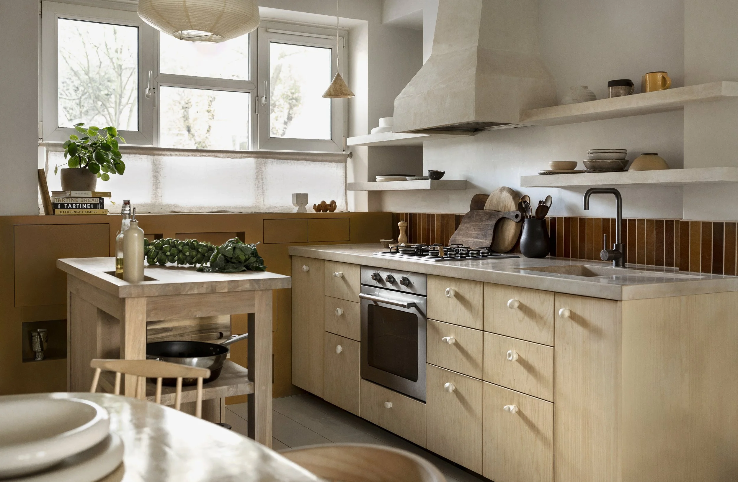 A cozy kitchen with light wood cabinets, a stainless steel oven, open shelves with dishes, a small dining table with a chair, and a window with sunlight. Decorative items include plants and books.
