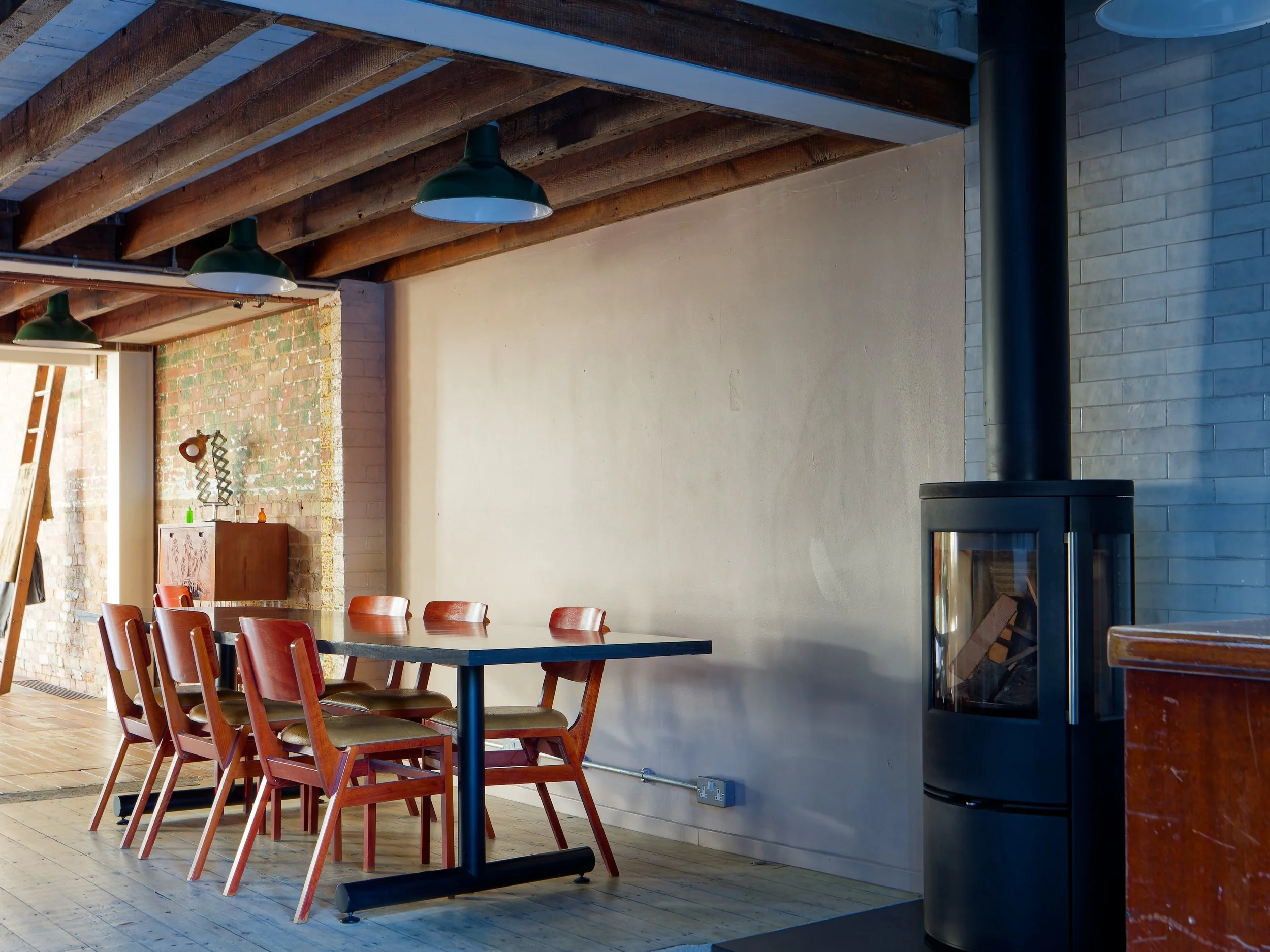 Dining area with wooden chairs around a table, exposed brick and painted walls, wood ceiling beams, and a black wood stove in a modern rustic interior.