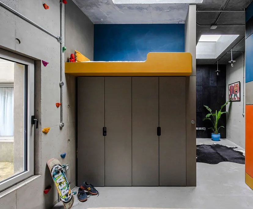 Indoor room with a climbing wall on the left, skateboard and shoes on the floor, cabinet with yellow bed above it, and a plant in a blue pot.