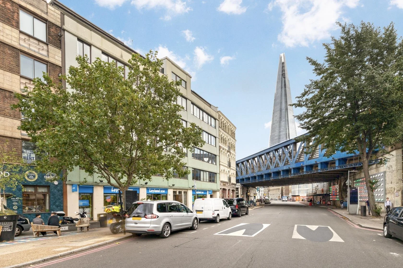 Street view of a city with cars parked along the curb, trees lining the sidewalk, and a blue elevated bridge leading to the skyscraper, The Shard, in the background under a partly cloudy sky.