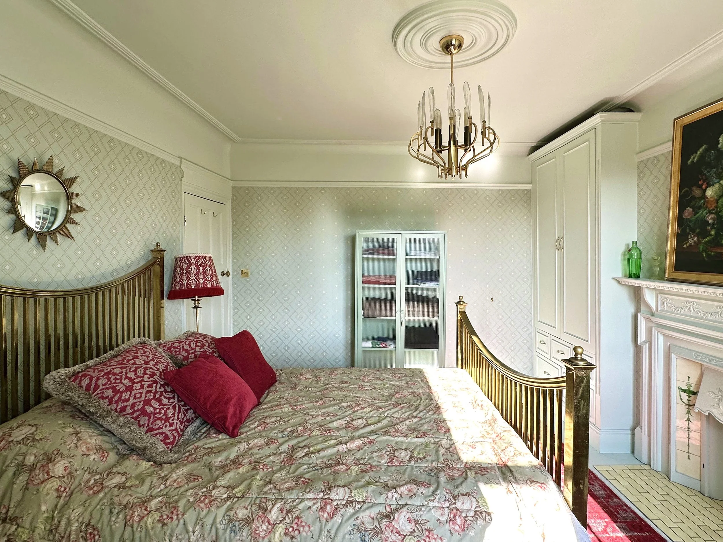 A bedroom with a bed covered in floral quilt and red pillows, a gold chandelier hanging from the ceiling, a white cabinet, a fireplace, and a mirror on the wall.
