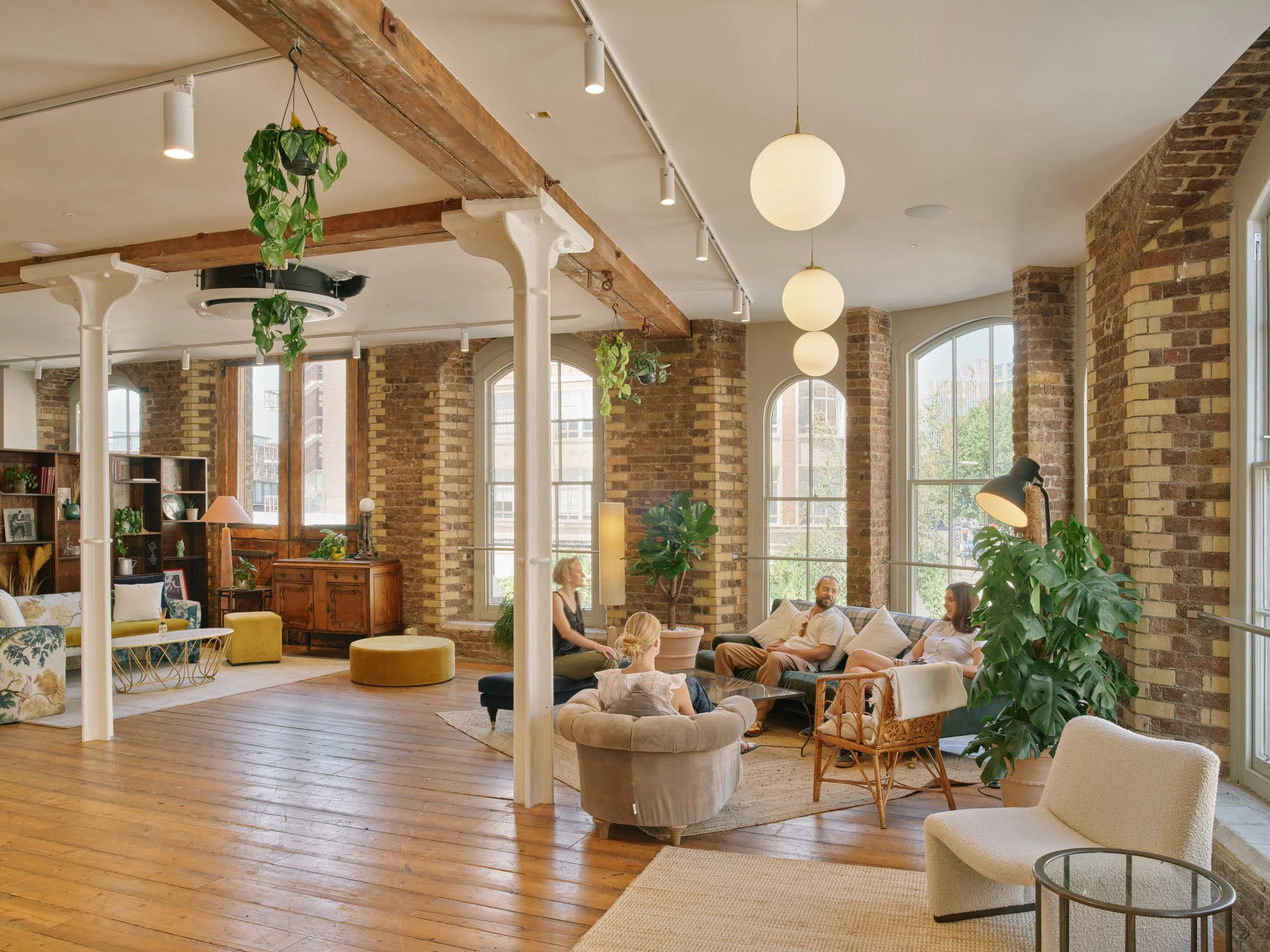 People sitting and chatting in a spacious, brightly lit room with large windows, brick walls, and wooden floors, decorated with plants and modern furniture.