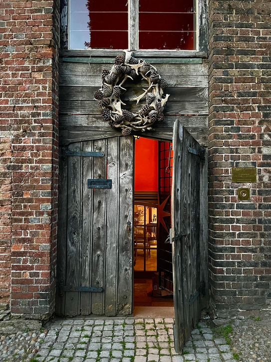 A rustic brick building with an open wooden door leading inside, decorated with a wreath made of pinecones and antlers, and a window above the door.