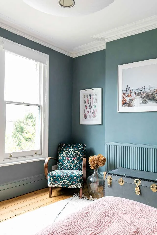 A corner of a bedroom with a blue armchair, a glass vase with dried flowers, a large grey storage trunk, and framed artwork on the blue wall. A window with white trim lets in natural light, and a pink quilted bedspread is partially visible in the for